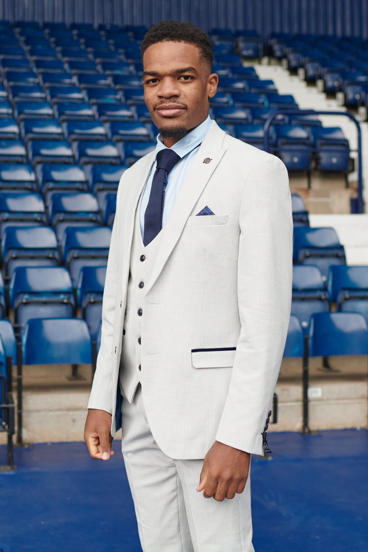 A man wearing the Marc Darcy WBA Collection - Bromley Suit, as worn by Grady Diangana, with a light gray jacket, blue shirt, and navy tie stands before empty blue stadium seats, looking at the camera with a slight smile.