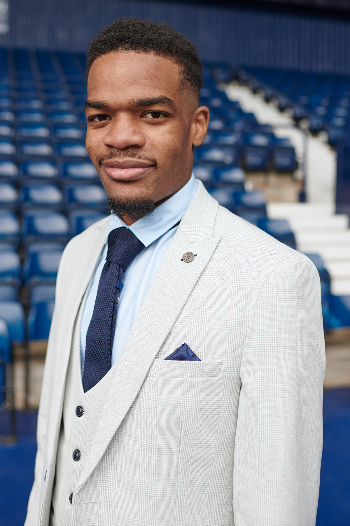 A man wears the Marc Darcy WBA Collection - Bromley Suit as worn by Grady Diangana, paired with a blue shirt and navy tie, standing and smiling in front of blue stadium seats.