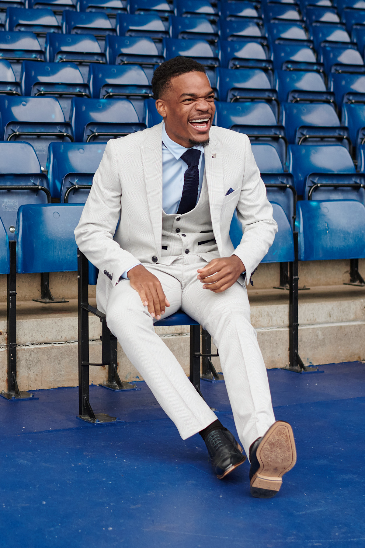 Grady Diangana laughs while seated in blue stadium seats, wearing the slim fit Bromley Suit from The WBA Collection by Marc Darcy.