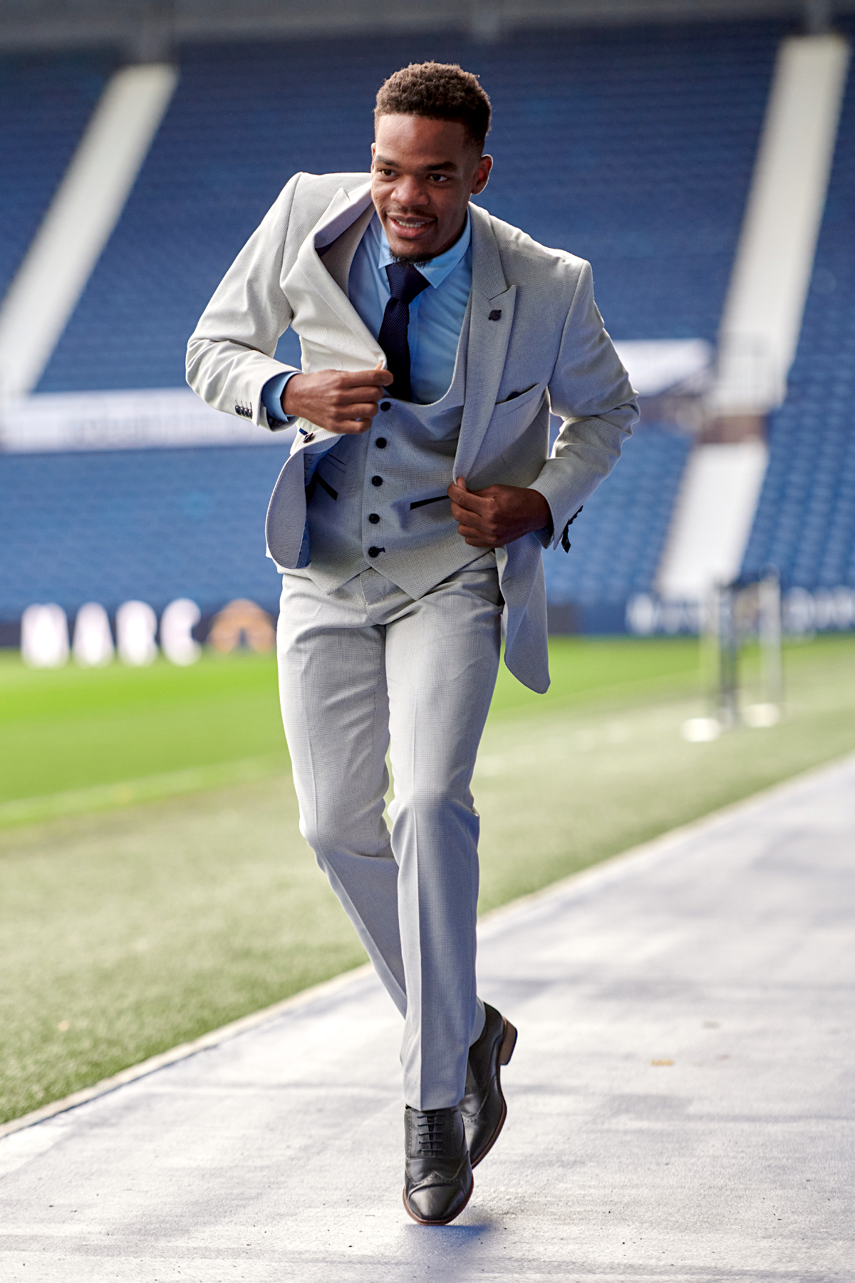 Grady Diangana walks on a stadium concourse adjusting his jacket, wearing the Marc Darcy WBA Collection Bromley Suit in slim fit light grey with a blue shirt and tie.