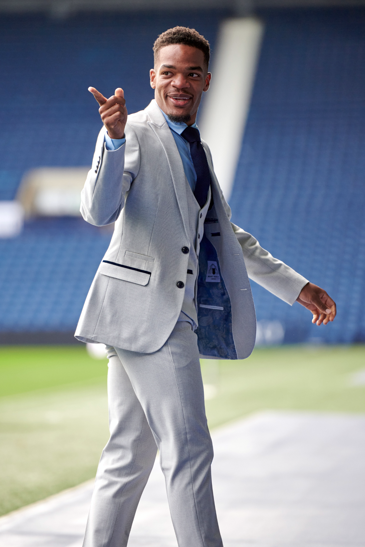 A man in the Marc Darcy WBA Collection - Bromley Suit as worn by Grady Diangana, pairs the slim fit, light grey suit with a blue shirt and smiles while walking by empty blue stadium seats.