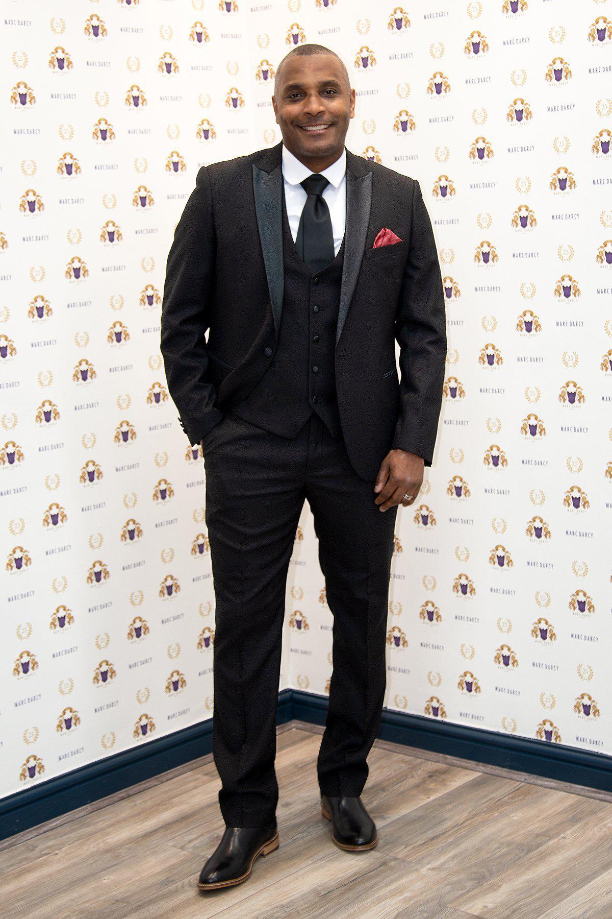 Former Crystal Palace footballer Clinton Morrison wears a marcdarcy DALTON Black Tux Lapel Suit with a red pocket square, smiling against a patterned backdrop with small logos and standing on a wood-paneled floor.