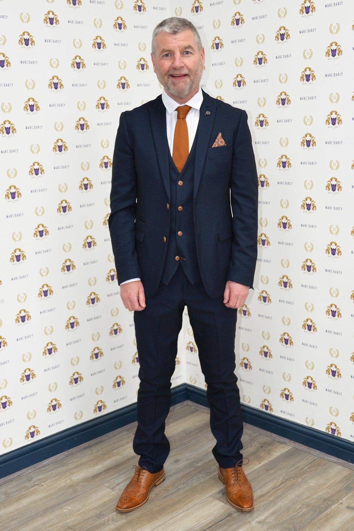 A man with gray hair and a beard stands on a wooden floor in front of a patterned backdrop, wearing the marcdarcy Manchester United Treble Winner Denis Irwin Max Navy Suit.