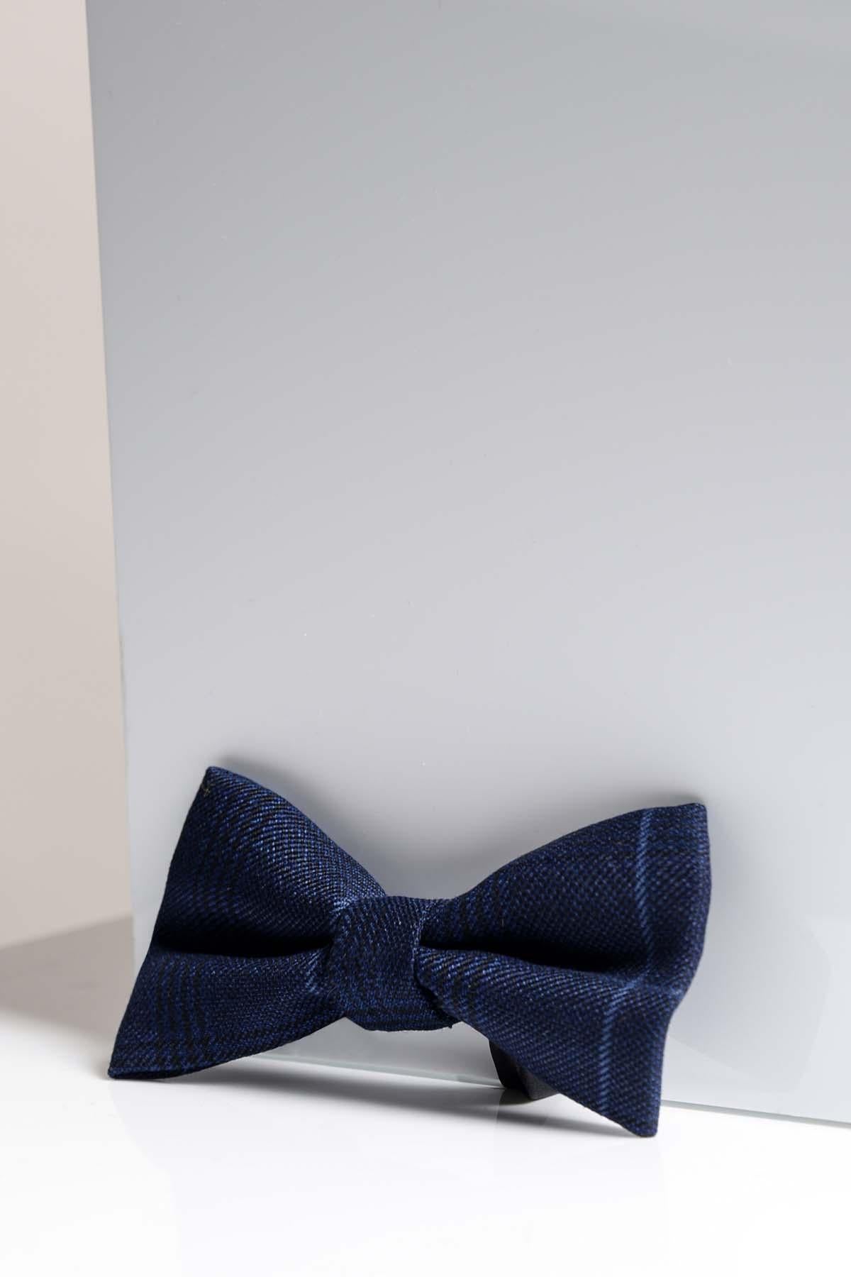 The EDINSON - Navy Sky Check Bow Tie by Accessory, pre-tied with an adjustable strap and subtle pattern, rests before a light gray reflective surface, its dark blue tone casting a gentle shadow underneath.