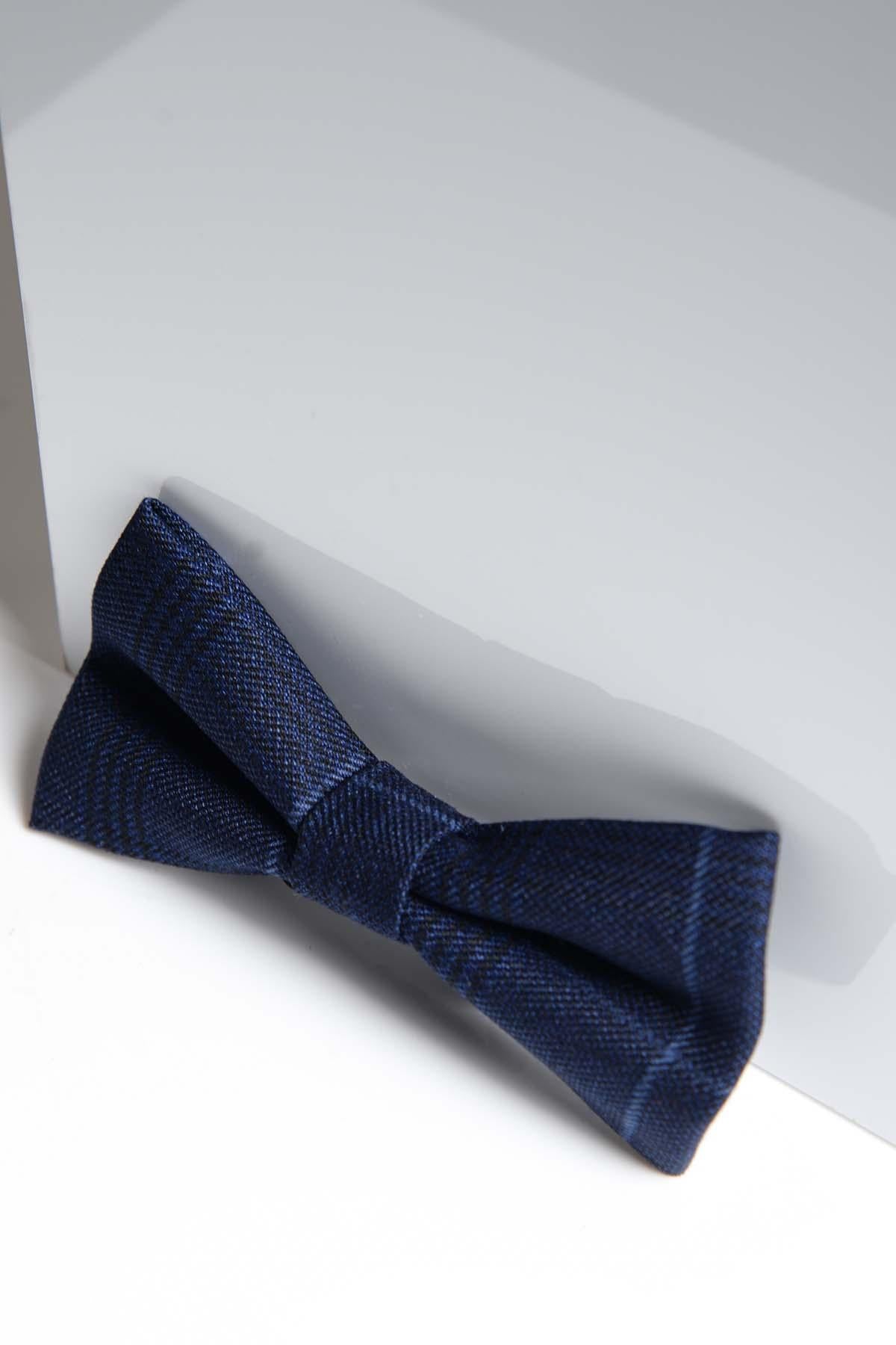 The EDINSON - Navy Sky Check Bow Tie by Accessory, featuring a textured finish, is displayed on a white glossy surface and leans against a reflective panel.