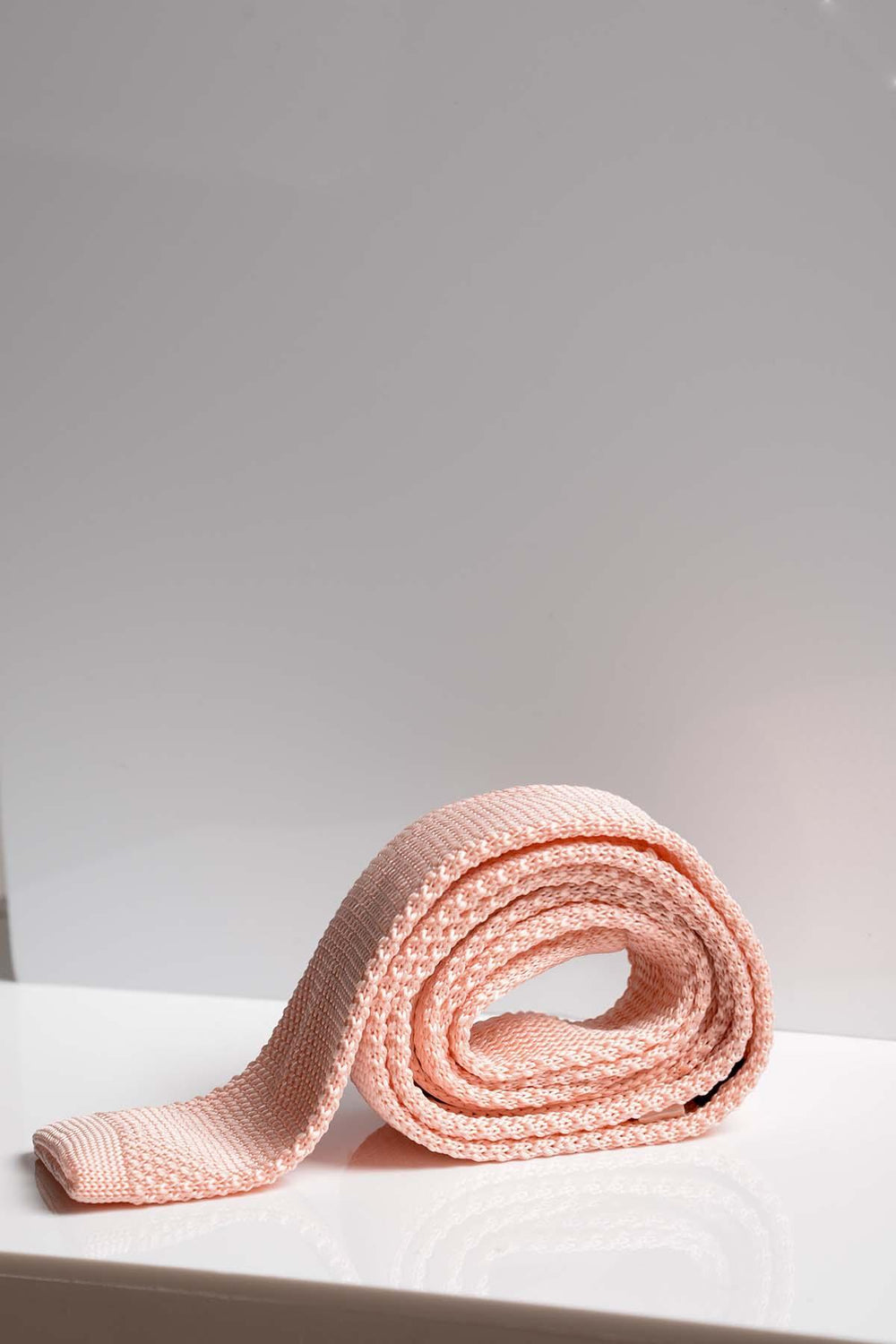 The Accessory KT - Peach Knitted Tie is rolled up on a white surface against a light grey background, making it an ideal summer wedding accessory.