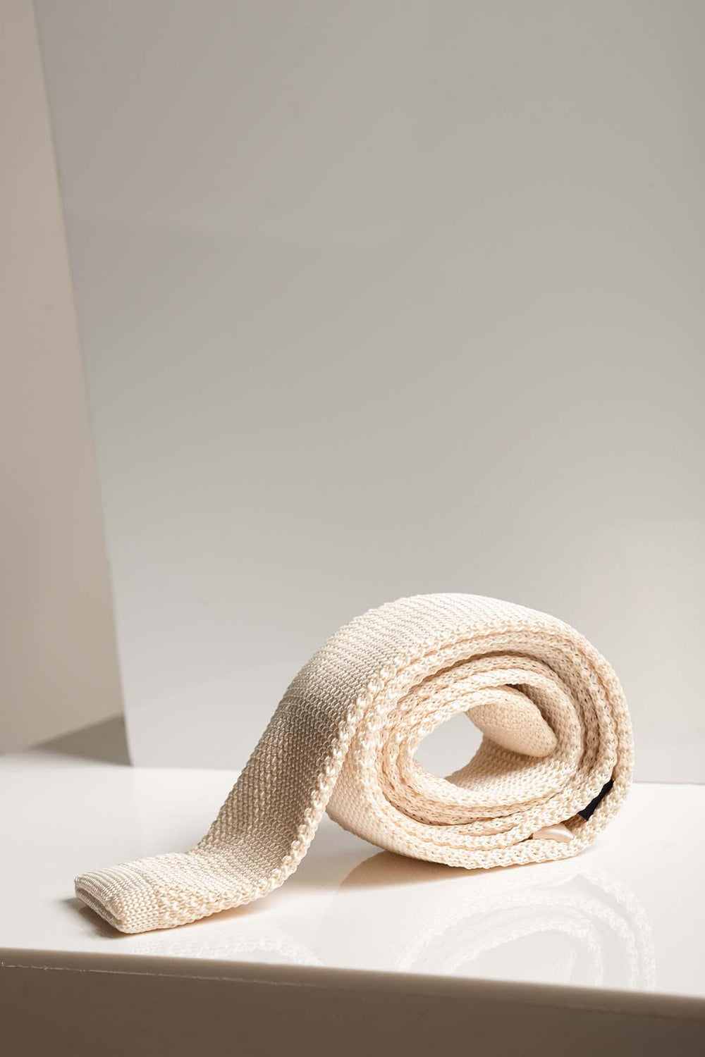The KT - Cream Knitted Tie by Accessory is rolled up and displayed on a glossy white surface with a neutral background, making it an elegant wedding accessory.
