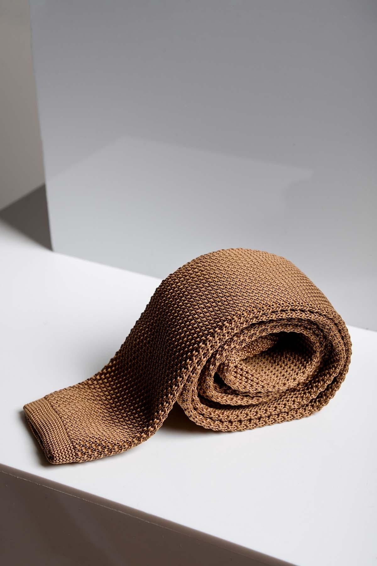 The KT - Dark Oak Knitted Tie by Accessory, with its textured design, is rolled up on a white surface against a gray and white background—a stylish choice for weddings.