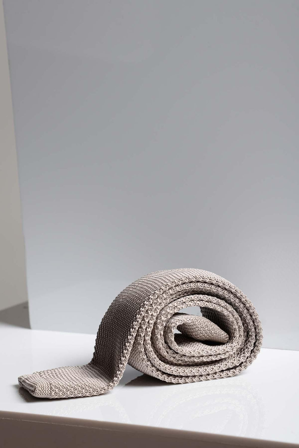The KT - Stone Knitted Tie by Accessory, in light grey with a modern textured design, is rolled up and displayed on a shiny white surface with a simple pale background.