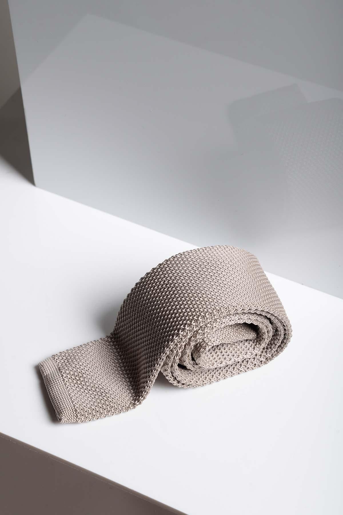 The KT - Stone Knitted Tie by Accessory, in light beige, is neatly rolled on a white surface, showcasing its modern textured design and casting a subtle shadow against the minimal backdrop.
