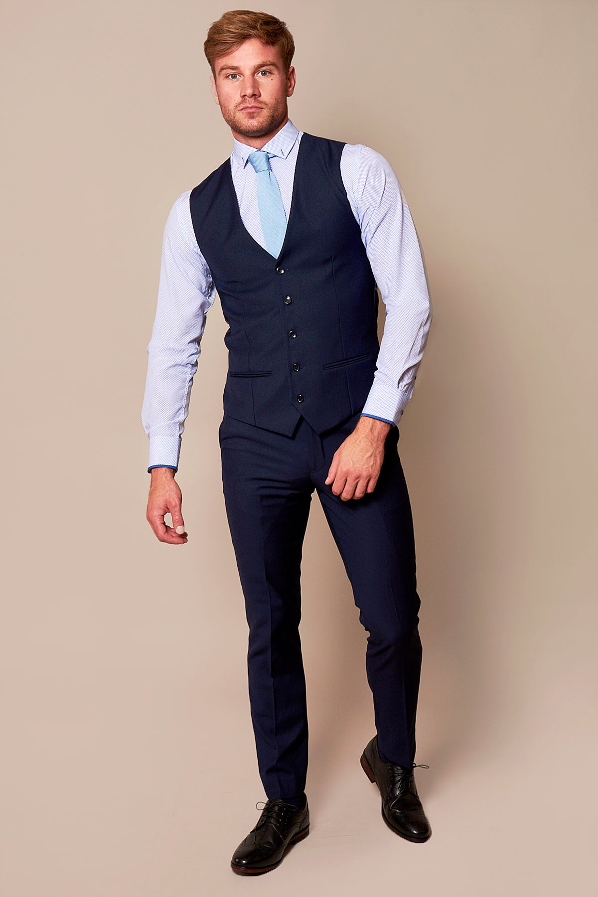 A man stands against a beige backdrop wearing the marcdarcy RAMBO Navy Formal Dress Waistcoat, styled with a matching navy suit, light blue shirt and tie, and black dress shoes. He gazes forward with a neutral expression.