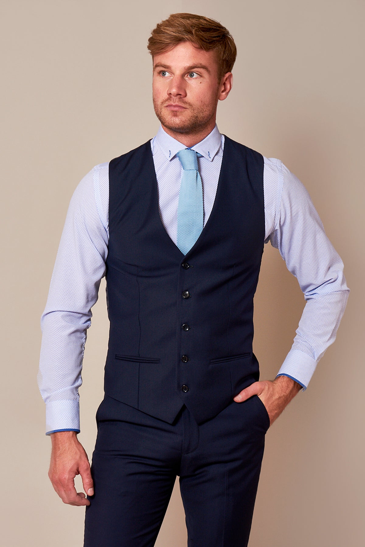 A man with light brown hair and a beard wears the marcdarcy RAMBO Navy Formal Dress Waistcoat with matching trousers, a light blue shirt, and blue tie, standing with one hand in his pocket against a beige background.