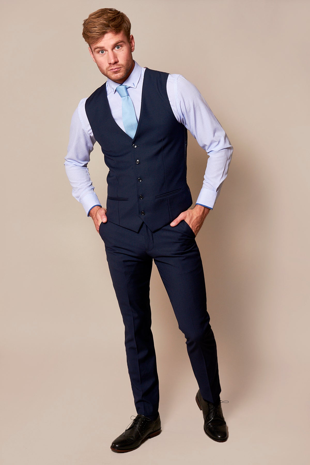 A man wears the marcdarcy RAMBO Navy Formal Dress Waistcoat with a matching navy suit, light blue shirt, tie, and black dress shoes, standing against a beige background with hands in his pockets and looking at the camera.