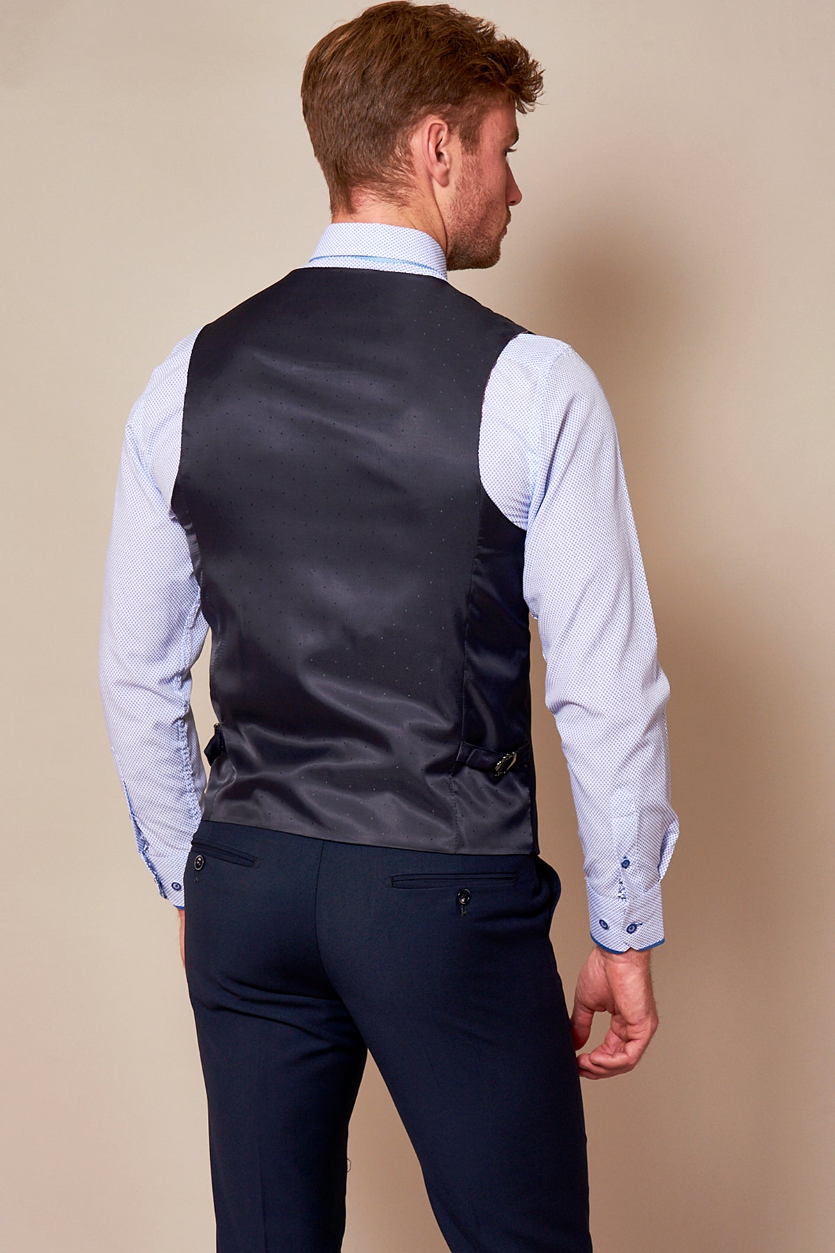 A man stands with his left hand relaxed by his side, wearing a light blue dress shirt, dark trousers, and the marcdarcy RAMBO Navy Formal Dress Waistcoat. He is seen from behind against a plain beige background.
