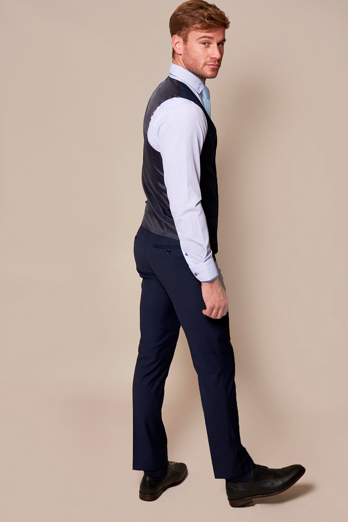 A man with light brown hair wears a marcdarcy RAMBO Navy Formal Dress Waistcoat, a light blue dress shirt, dark blue pants, and black shoes. He stands backward looking over his shoulder against a plain beige background.