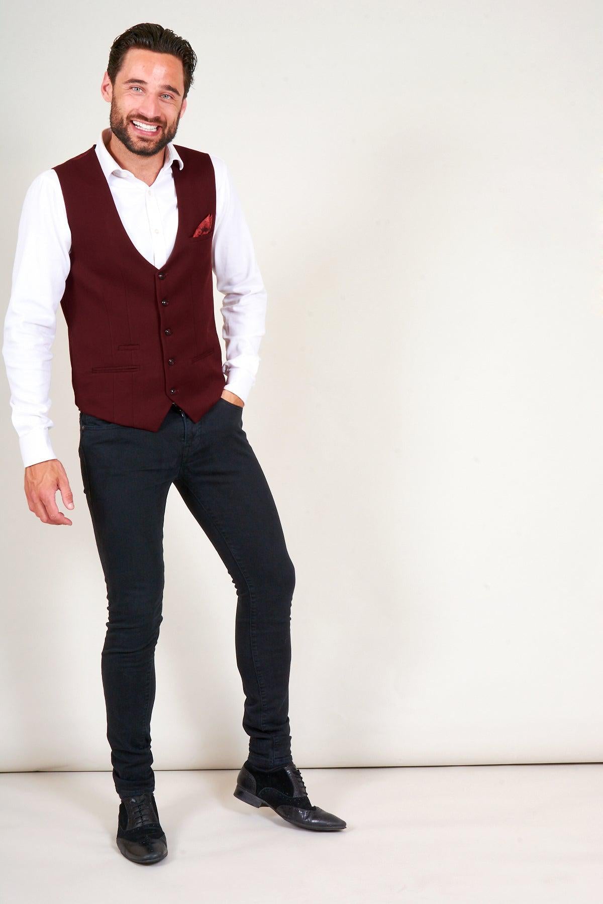 A smiling man in a white shirt, marcdarcy's KELLY Wine Single Breasted Waistcoat, black jeans, and black shoes stands with his hand in his pocket against a plain background.