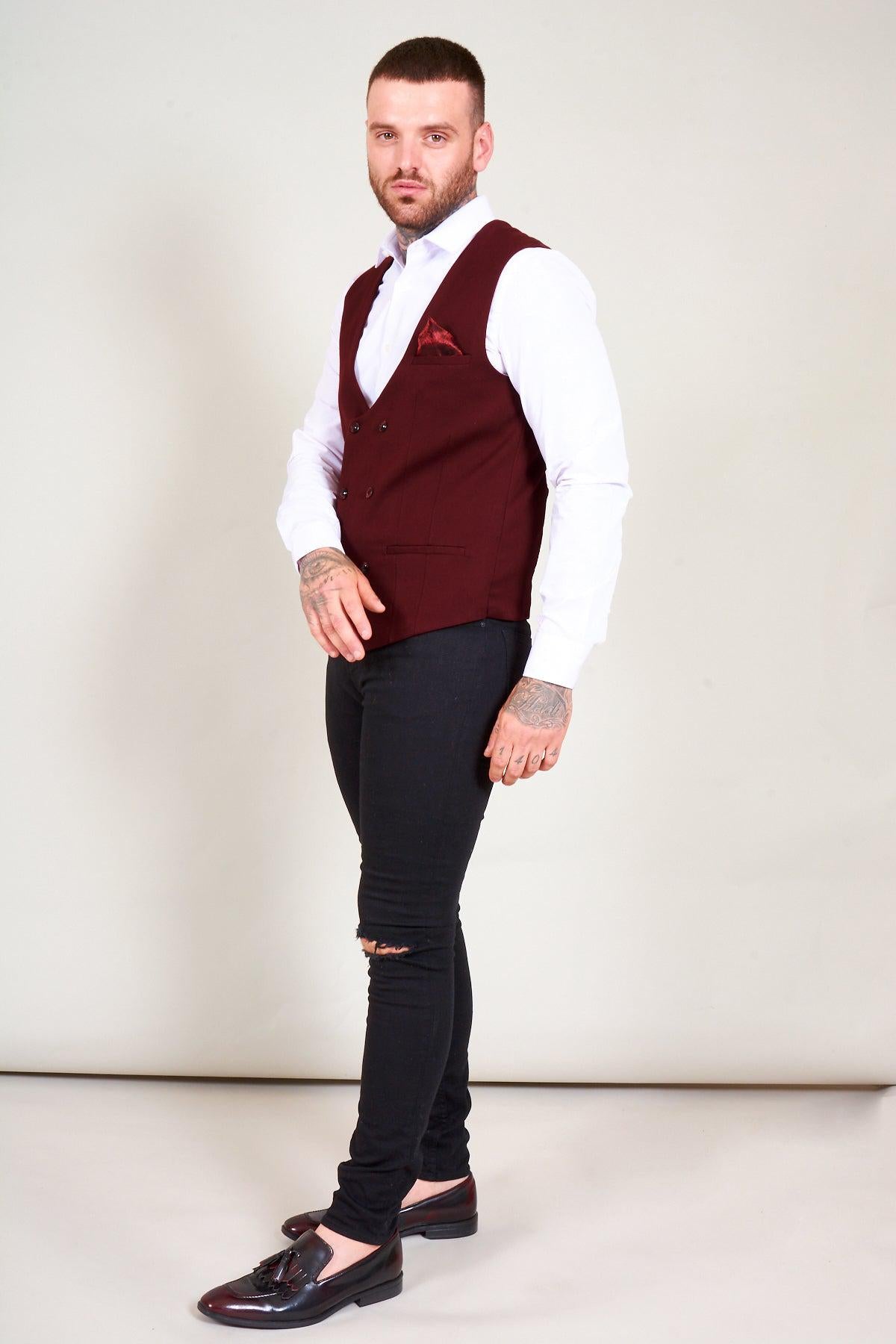 A man with short hair and a beard stands sideways in a studio, wearing the marcdarcy KELLY Wine Double Breasted Waistcoat over a white shirt, paired with black ripped jeans and black loafers, looking toward the camera.