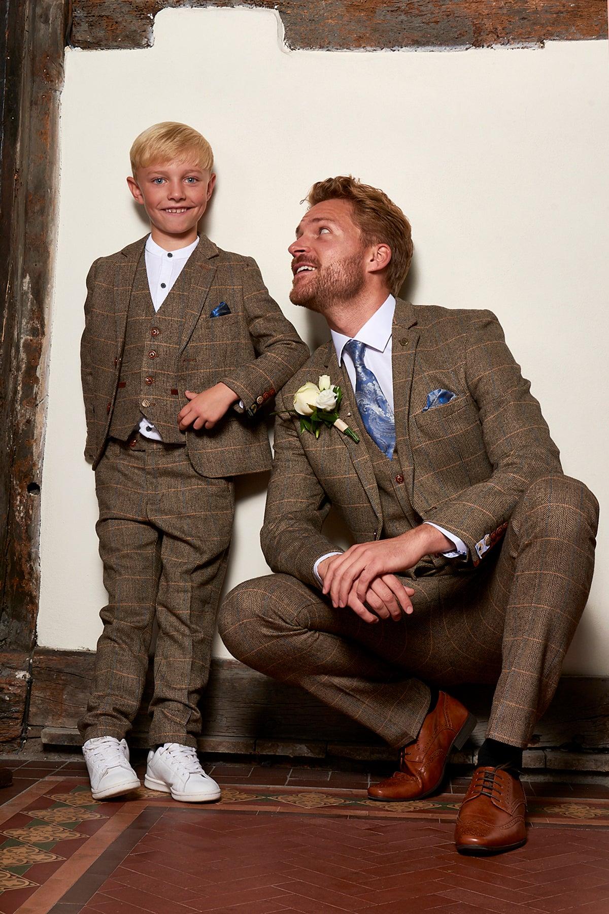 A man and young boy, both in marcdarcy Matching Father & Son Men’s TED Tan Tweed Check Three Piece Suits, smile together. The man wears a blue tie and boutonniere as they pose by a rustic wall with tiled flooring.
