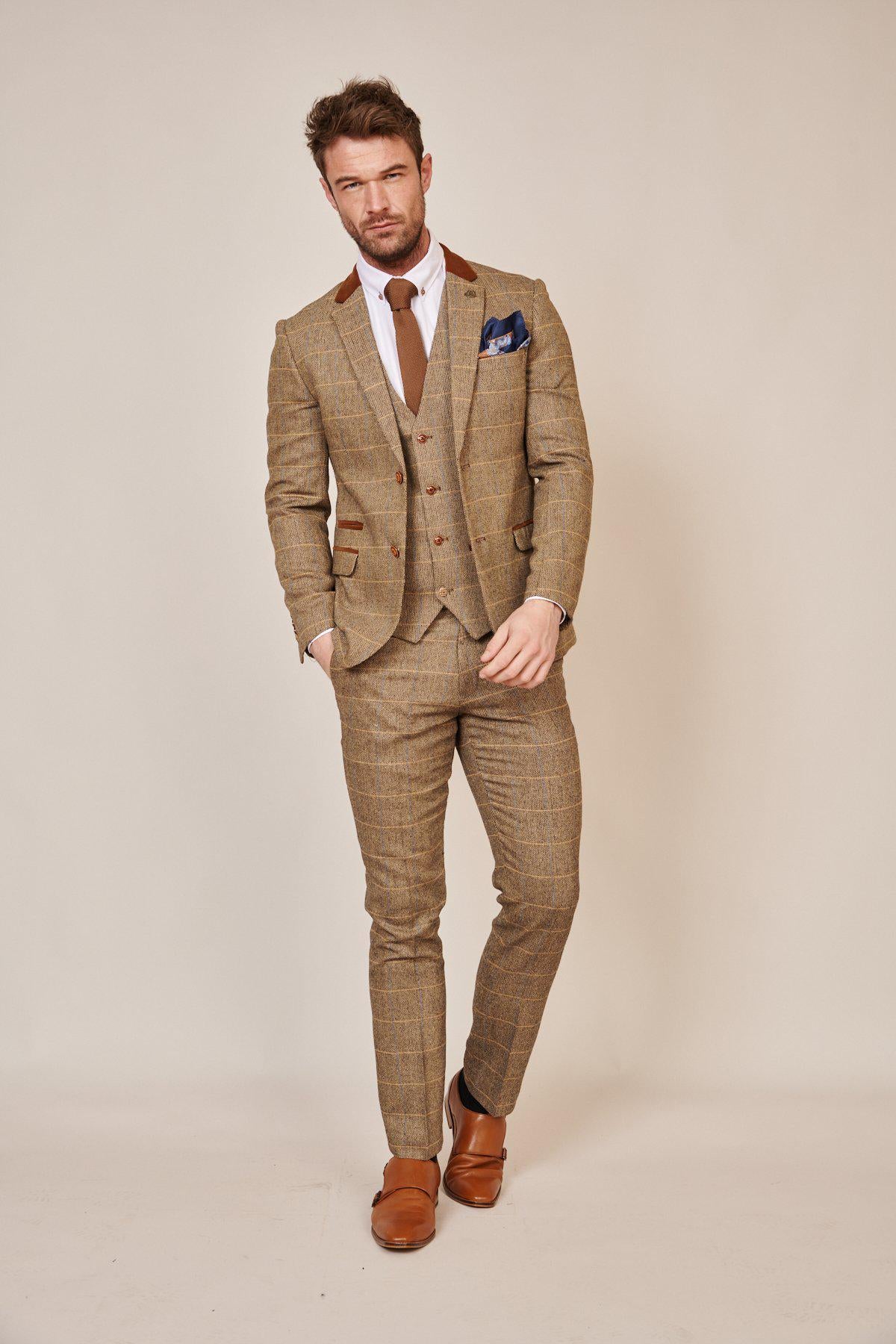 A man stands against a beige background wearing the marcdarcy Blogger Style | DX7 Suit as worn by Jonny Hammond, paired with a white shirt, brown tie and shoes, and a blue pocket square, with one hand in his pocket.