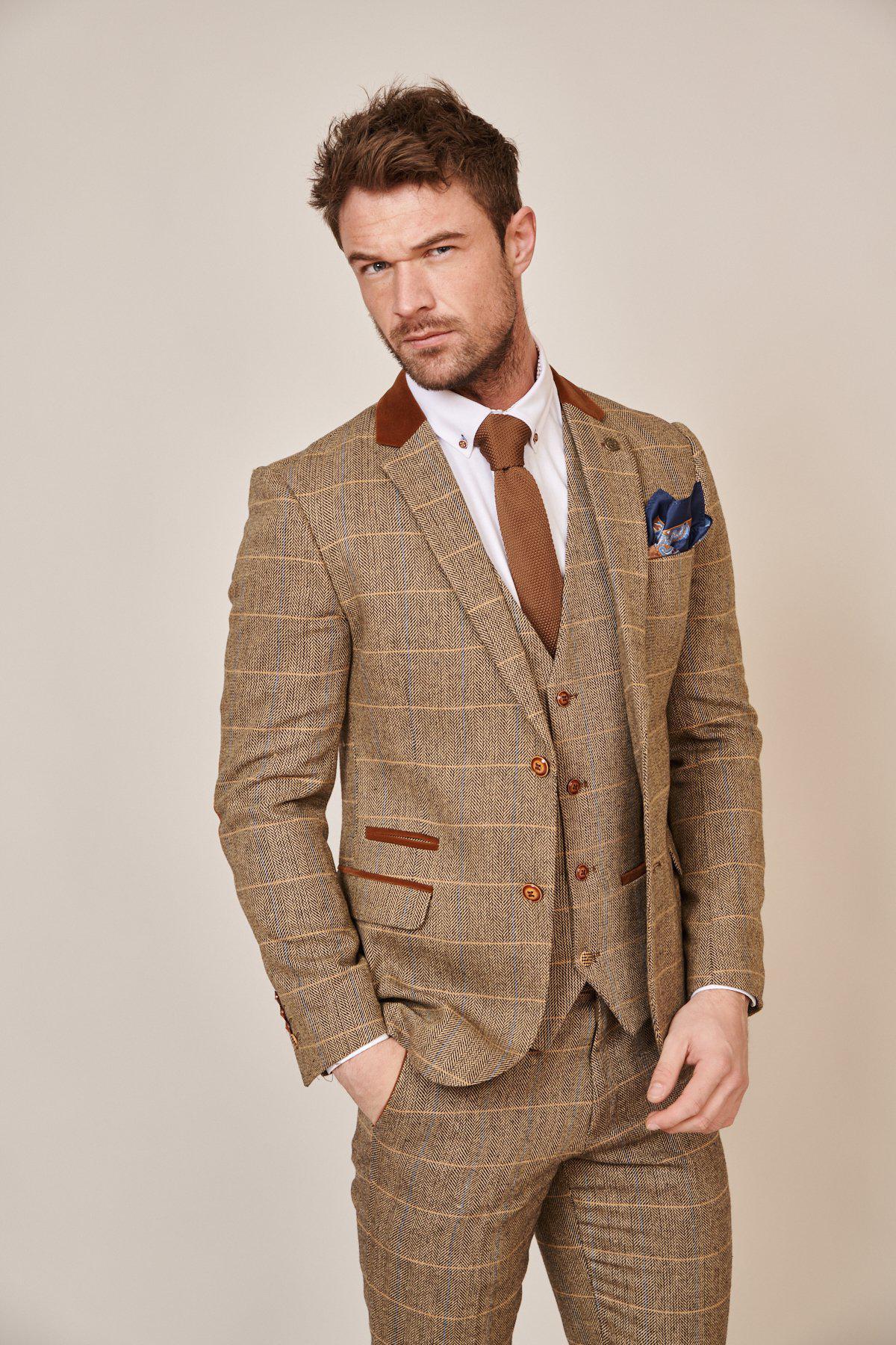 A man stands confidently against a plain light background, wearing the marcdarcy Blogger Style | DX7 Suit as worn by Jonny Hammond—a brown herringbone slim fit three-piece paired with a white shirt, brown tie, and blue pocket square.
