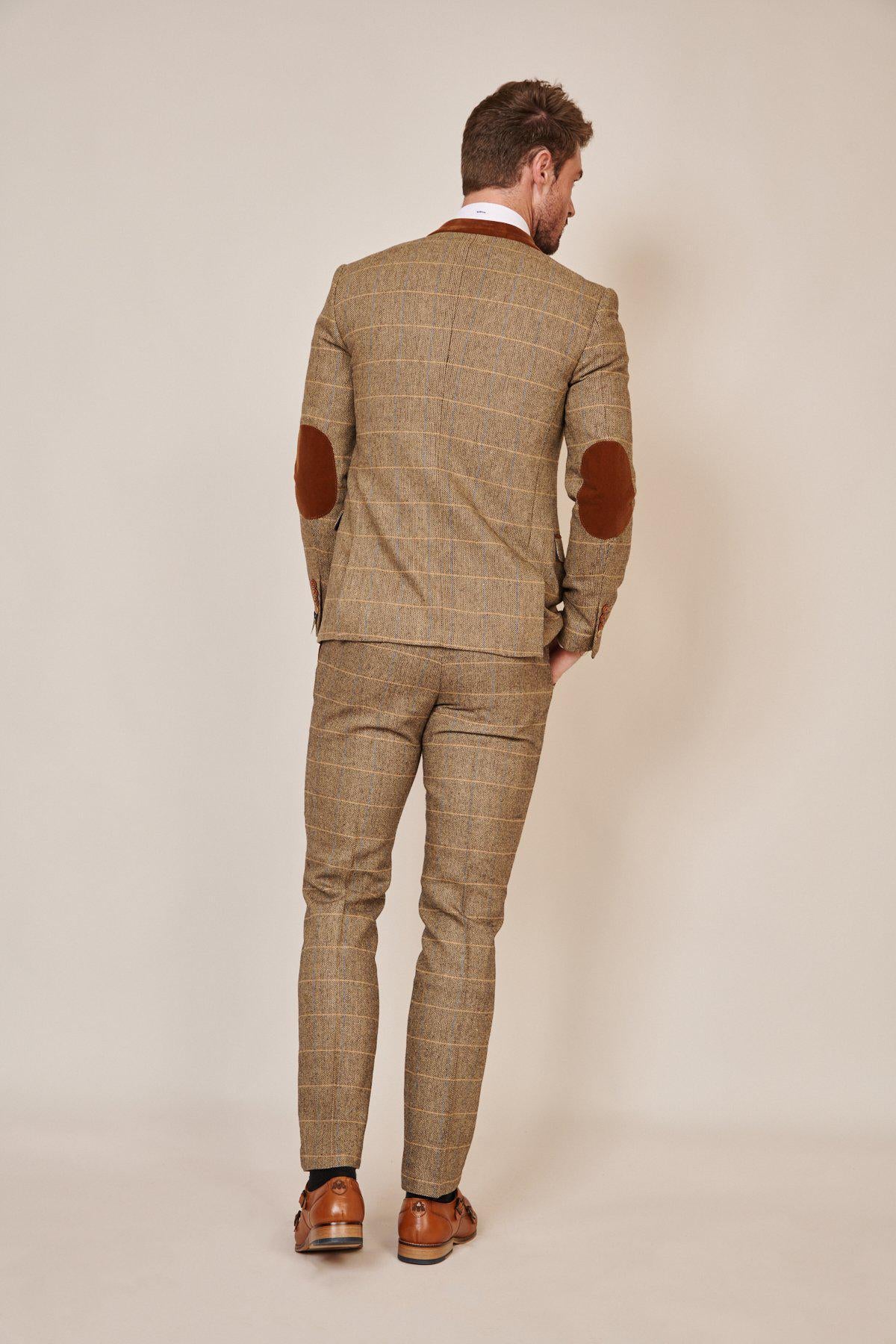 A man stands facing away, wearing the marcdarcy Blogger Style | DX7 Suit as worn by Jonny Hammond, featuring a brown herringbone pattern with brown elbow patches, complemented by brown dress shoes and a slim fit blazer.