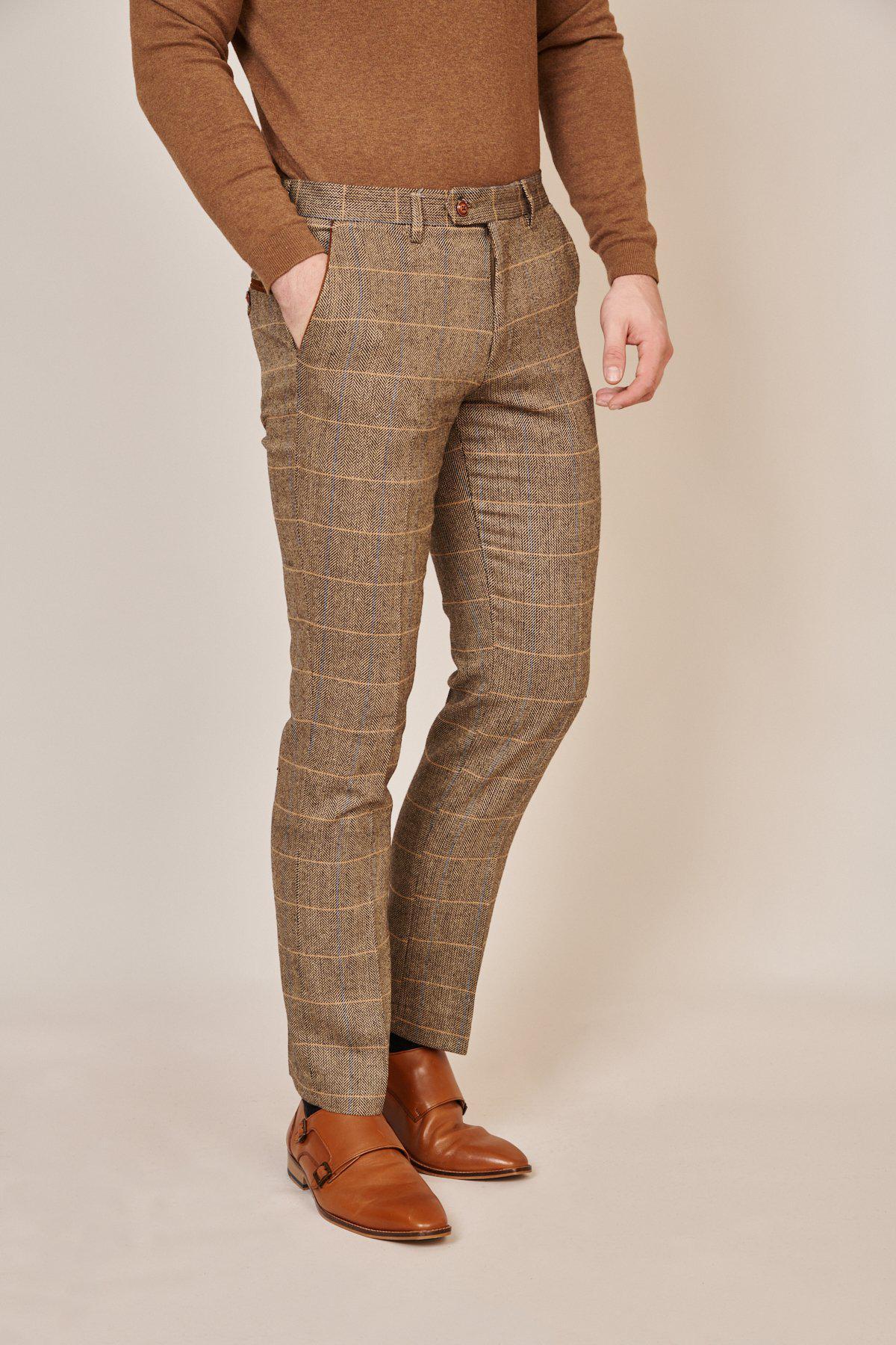 A man wears the marcdarcy Blogger Style | DX7 Suit As Worn By Jonny Hammond, featuring a slim fit blazer with a velvet contrast collar, brown crew-neck sweater, plaid trousers, and double monk strap shoes against a beige background.