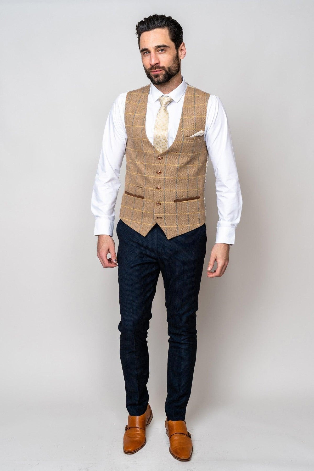 A bearded man wears the marcdarcy DX7 - Oak Tweed Check Waistcoat over a white shirt with a tan patterned tie, paired with navy pants and tan monk strap shoes.