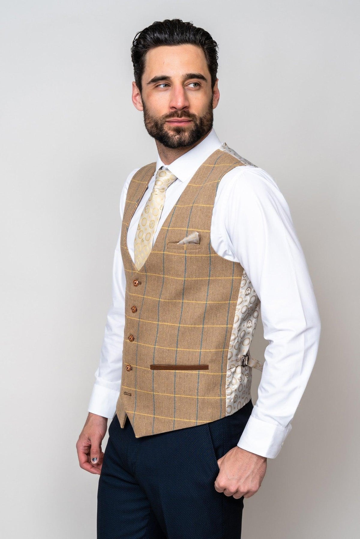 A man with dark hair and beard wears a marcdarcy DX7 - Oak Tweed Check Waistcoat, white dress shirt, light patterned tie, navy trousers, and beige pocket square, standing against a plain light background and looking to the side.