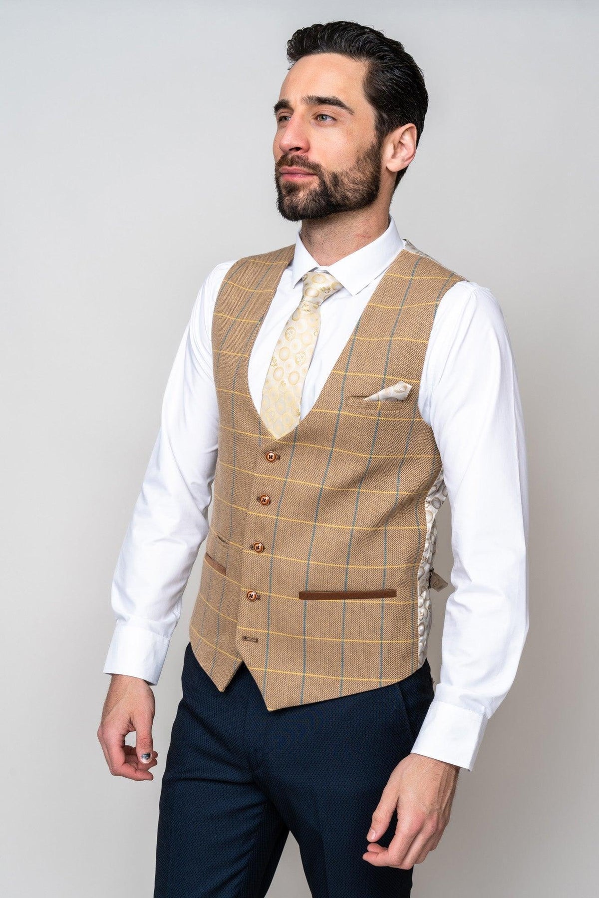 A man with dark hair and a beard wears the marcdarcy DX7 - Oak Tweed Check Waistcoat over a white shirt, cream patterned tie, navy trousers, and pocket square, standing against a plain light background.