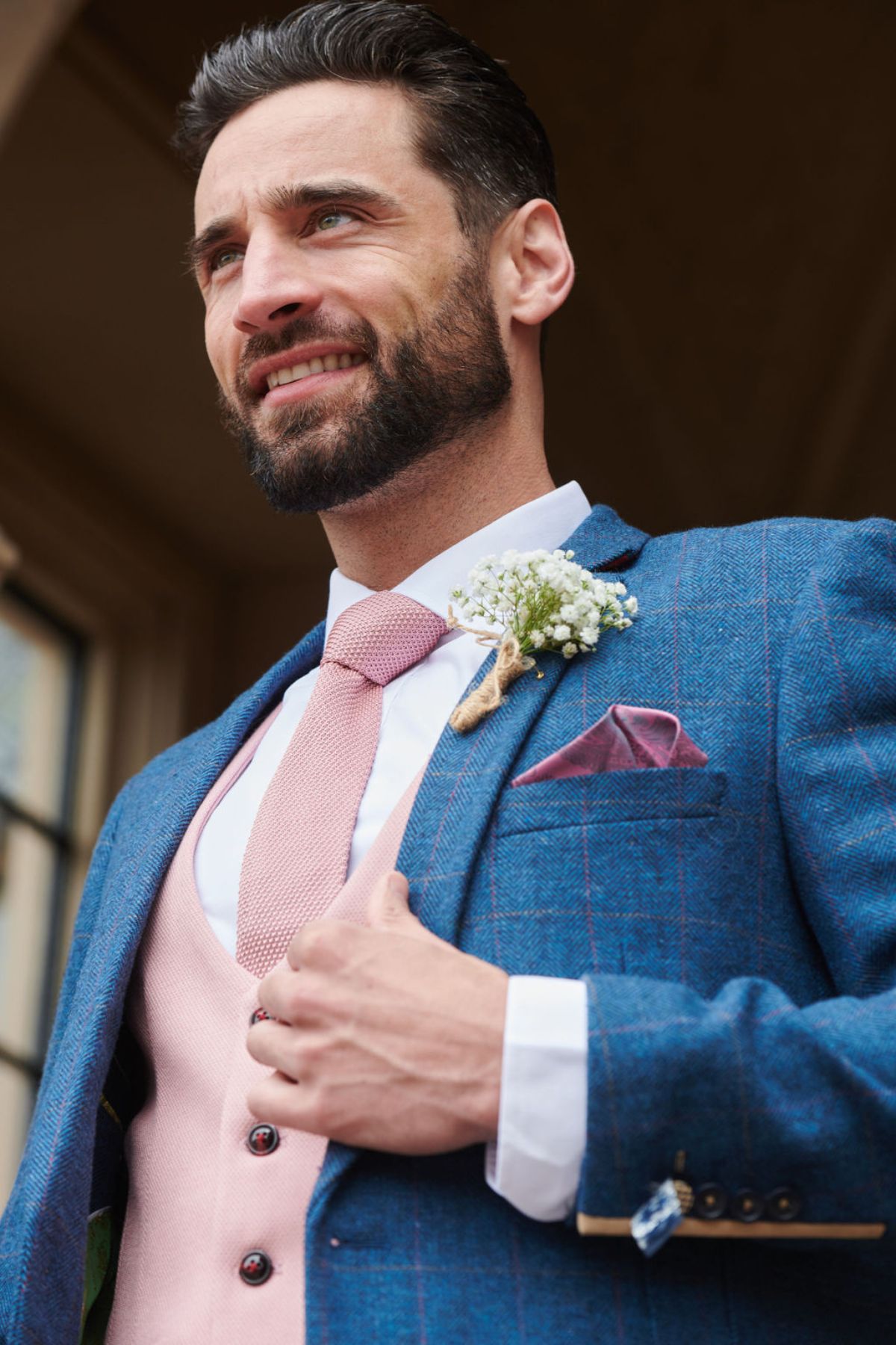 A groom smiles as he adjusts his marcdarcy DION blue tweed check suit with a Kelvin pink waistcoat and tie, finished with a baby's breath boutonnière.