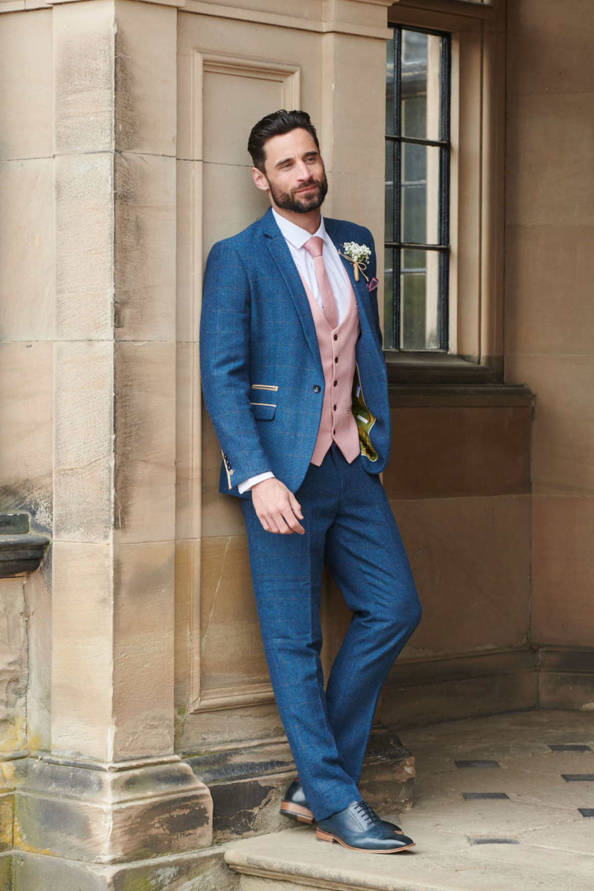 A bearded man leans against a stone building, wearing the marcdarcy DION Blue Tweed Check Suit with Kelvin Pink Waistcoat, complete with boutonniere and pocket square—showcasing stylish, mix-and-match wedding attire near a window.