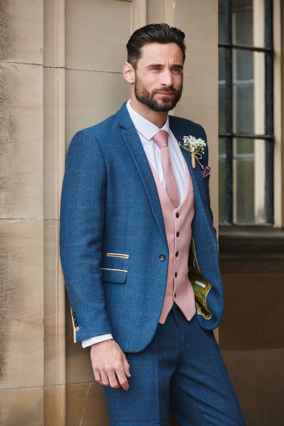 A man with dark hair and a beard stands by a stone wall, wearing the marcdarcy DION Blue Tweed Check Suit with a Kelvin Pink Waistcoat, pink tie, white shirt, and boutonnière, in front of a large window.