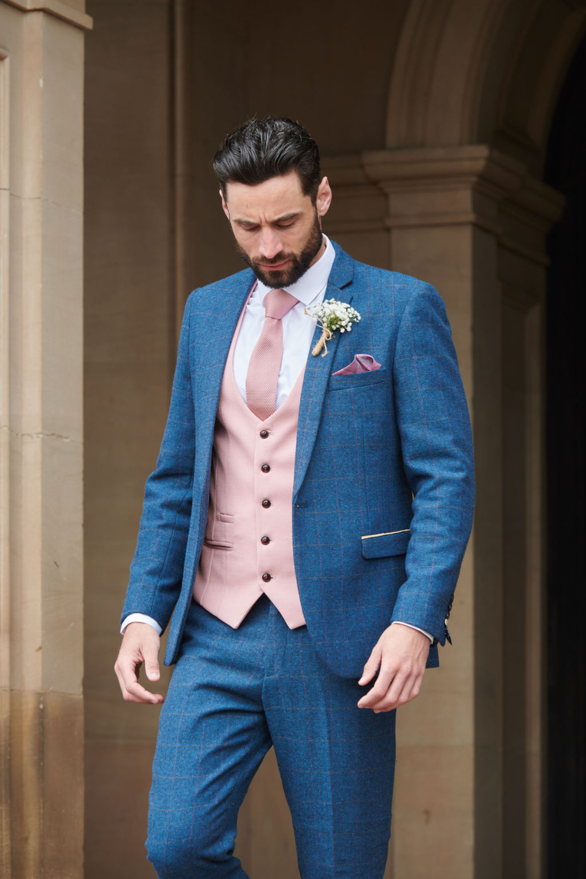 A man in the marcdarcy DION blue tweed check suit with Kelvin pink waistcoat stands outdoors by a stone building, boutonnière on his lapel, looking down with a serious expression—a modern mix-and-match wedding look.