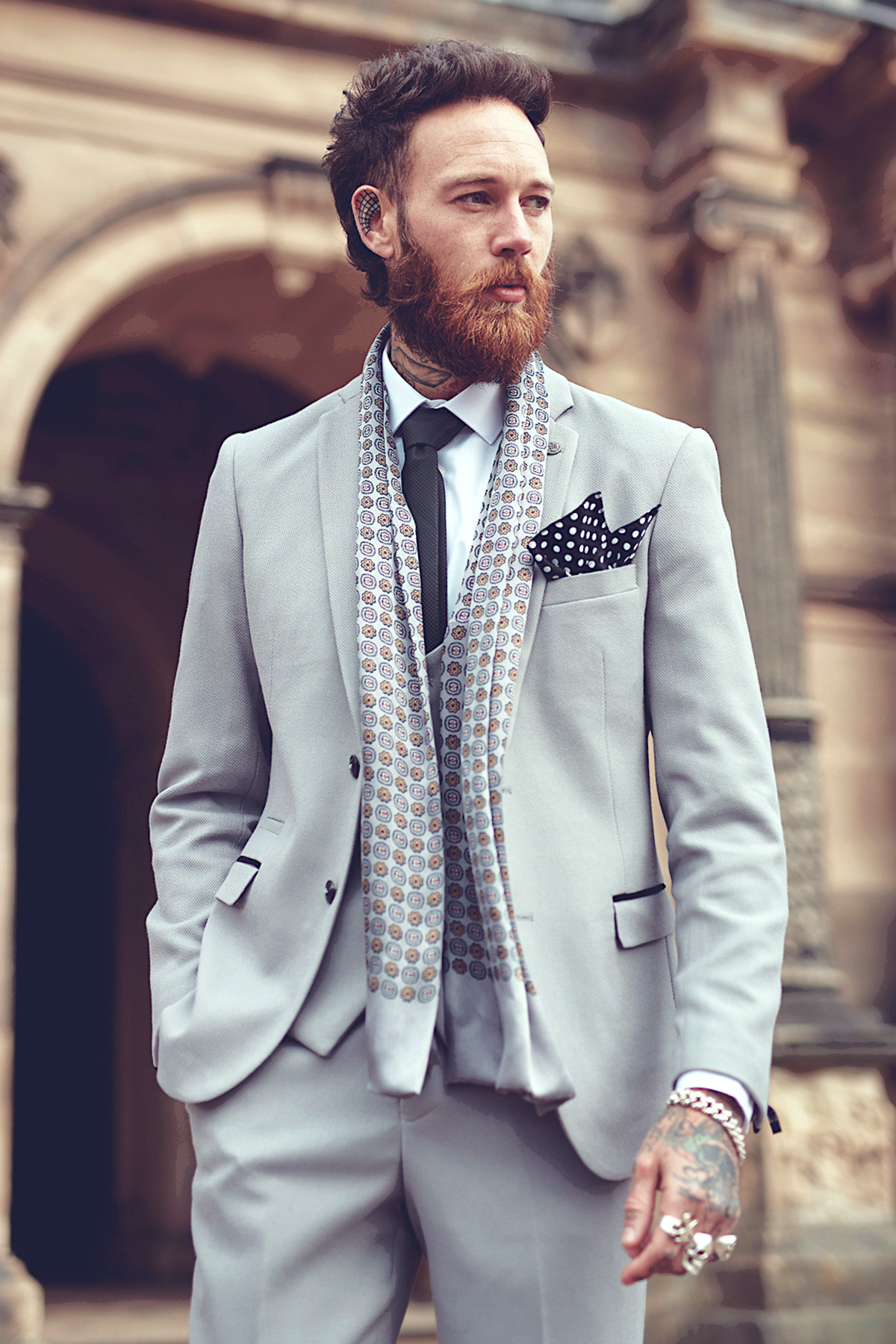 A bearded, tattooed man wearing the marcdarcy EDWIN Silver Grey Notch Lapel Three Piece Suit stands outdoors by an ornate stone building, sporting a patterned scarf, polka dot pocket square, and black tie with his hands in his pockets.