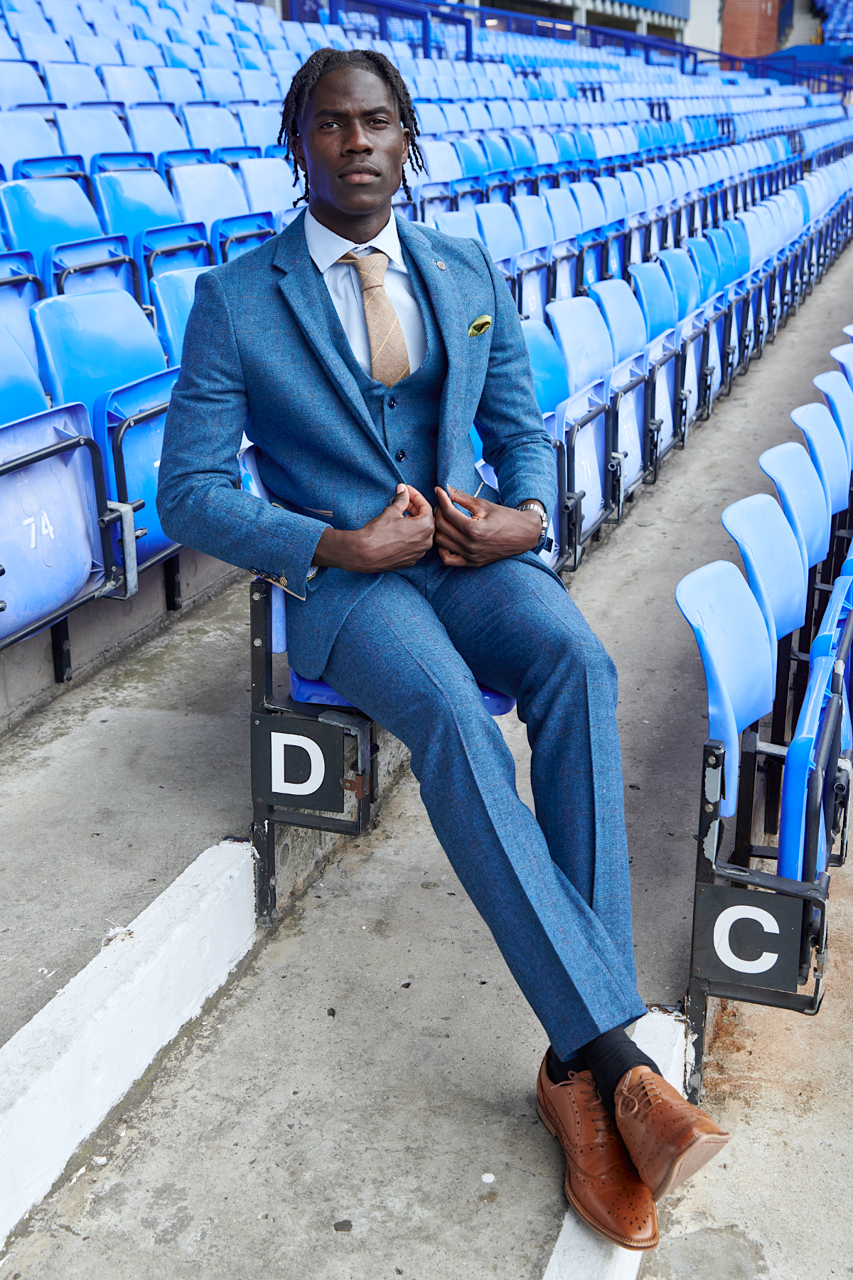 Amadou Onana sits confidently on a stadium seat in marcdarcy's The Everton Collection | Dion Blue suit, surrounded by empty blue chairs, elegantly showcasing refined men’s formalwear.