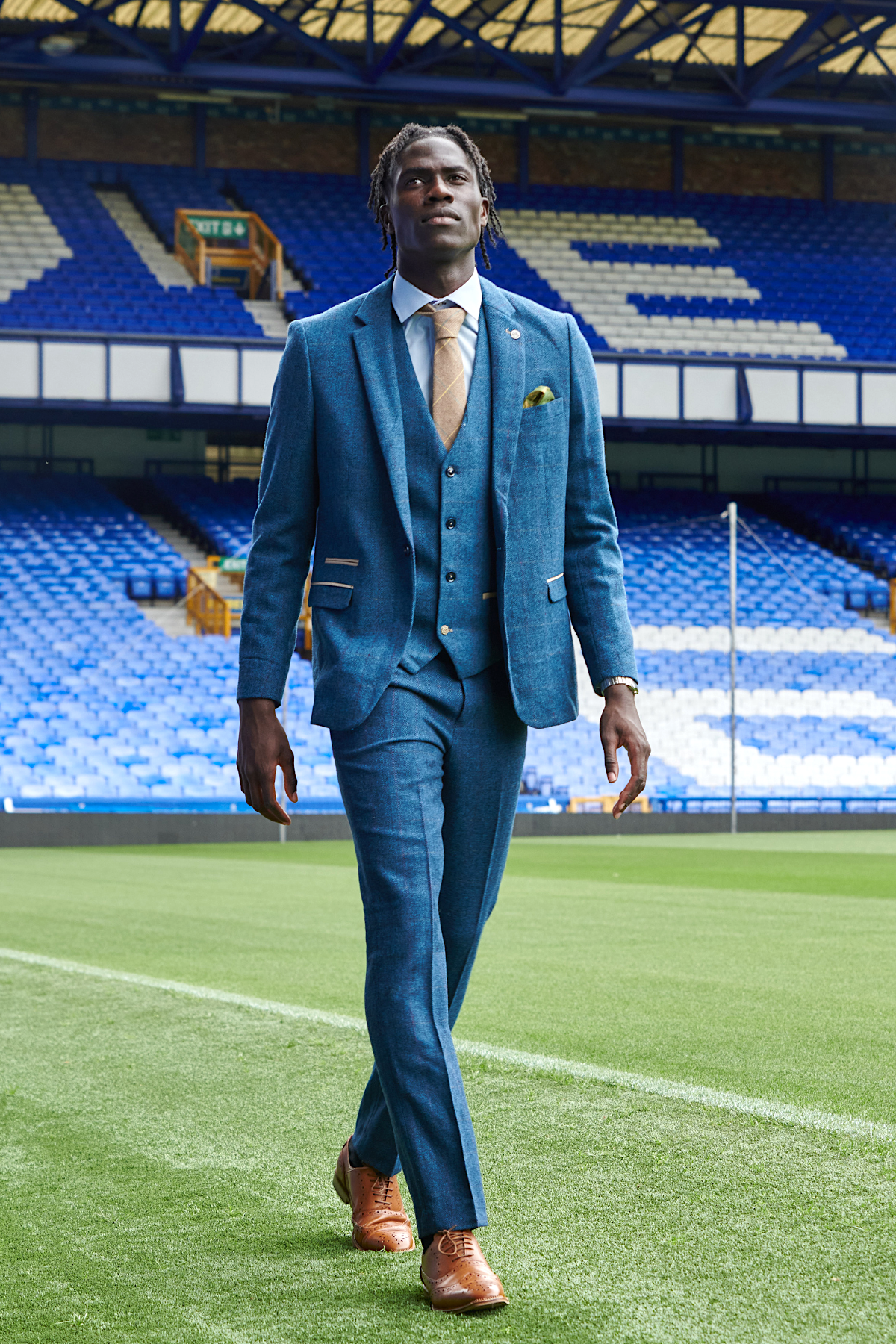 Amadou Onana wears The Everton Collection Dion Blue suit by marcdarcy, confidently walking on a football field with empty blue stadium seats and stands in the background.