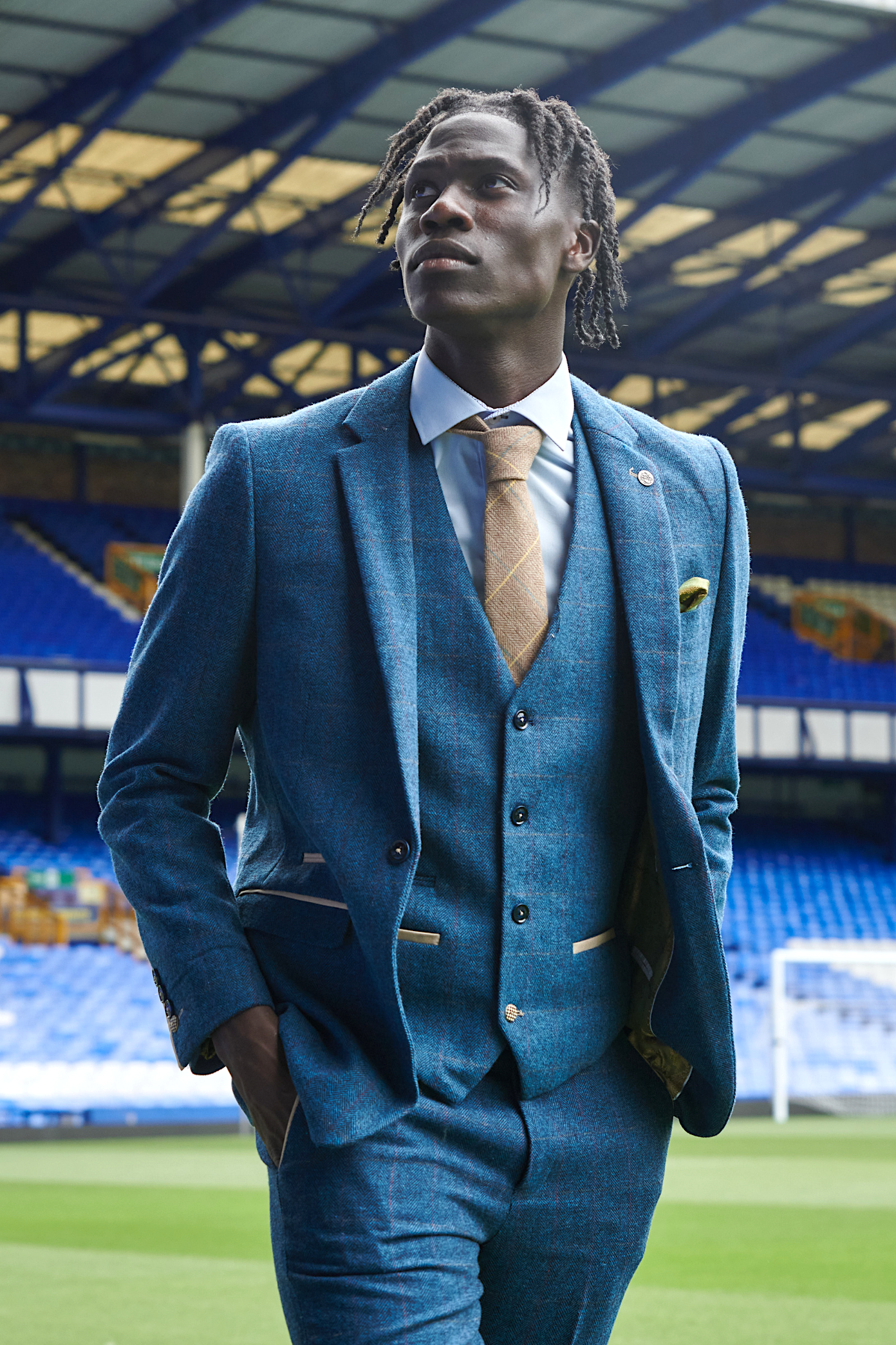 Amadou Onana models marcdarcy’s The Everton Collection in Dion Blue, standing on a football field with empty stadium seats and a goalpost behind him, his formal look contrasting with the sporty setting.
