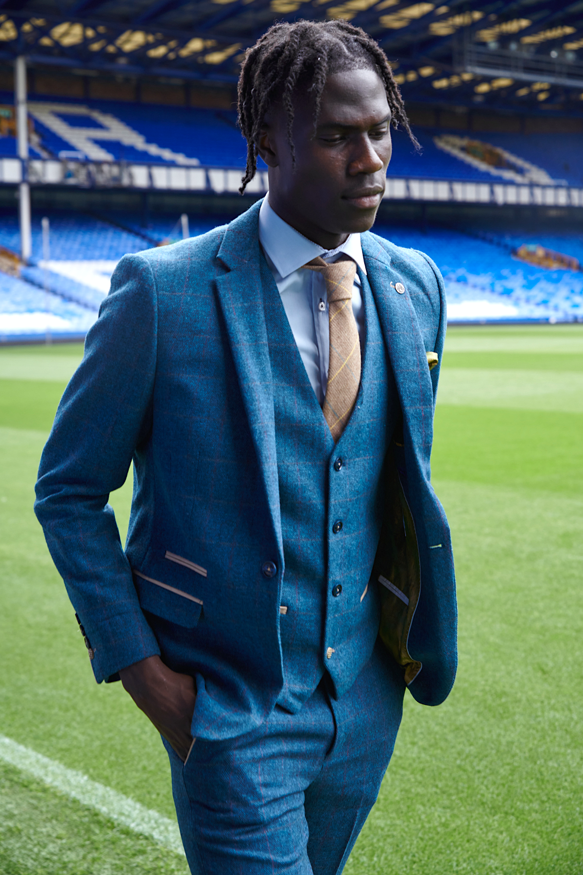 Amadou Onana wears a marcdarcy Dion Blue suit from The Everton Collection, walking across a football stadium field with blue seats in the background, looking down with one hand in his pocket.