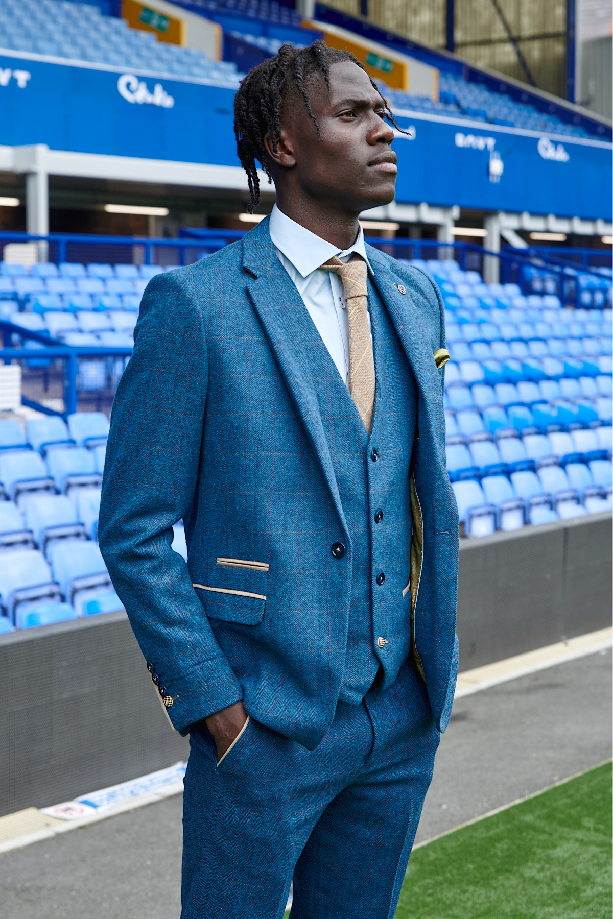 Amadou Onana wears The Everton Collection Dion Blue suit by marcdarcy—styled with a light shirt and tan tie—as he stands confidently before empty blue stadium seats, highlighting men's formal wear.