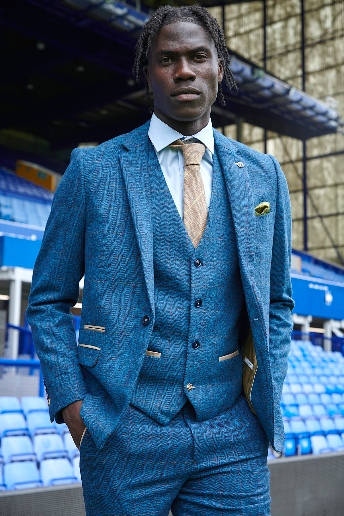 Amadou Onana stands confidently in a stadium, wearing marcdarcy’s Dion Blue suit from The Everton Collection—a bold display of men's formal wear amidst blue seats and stadium architecture.