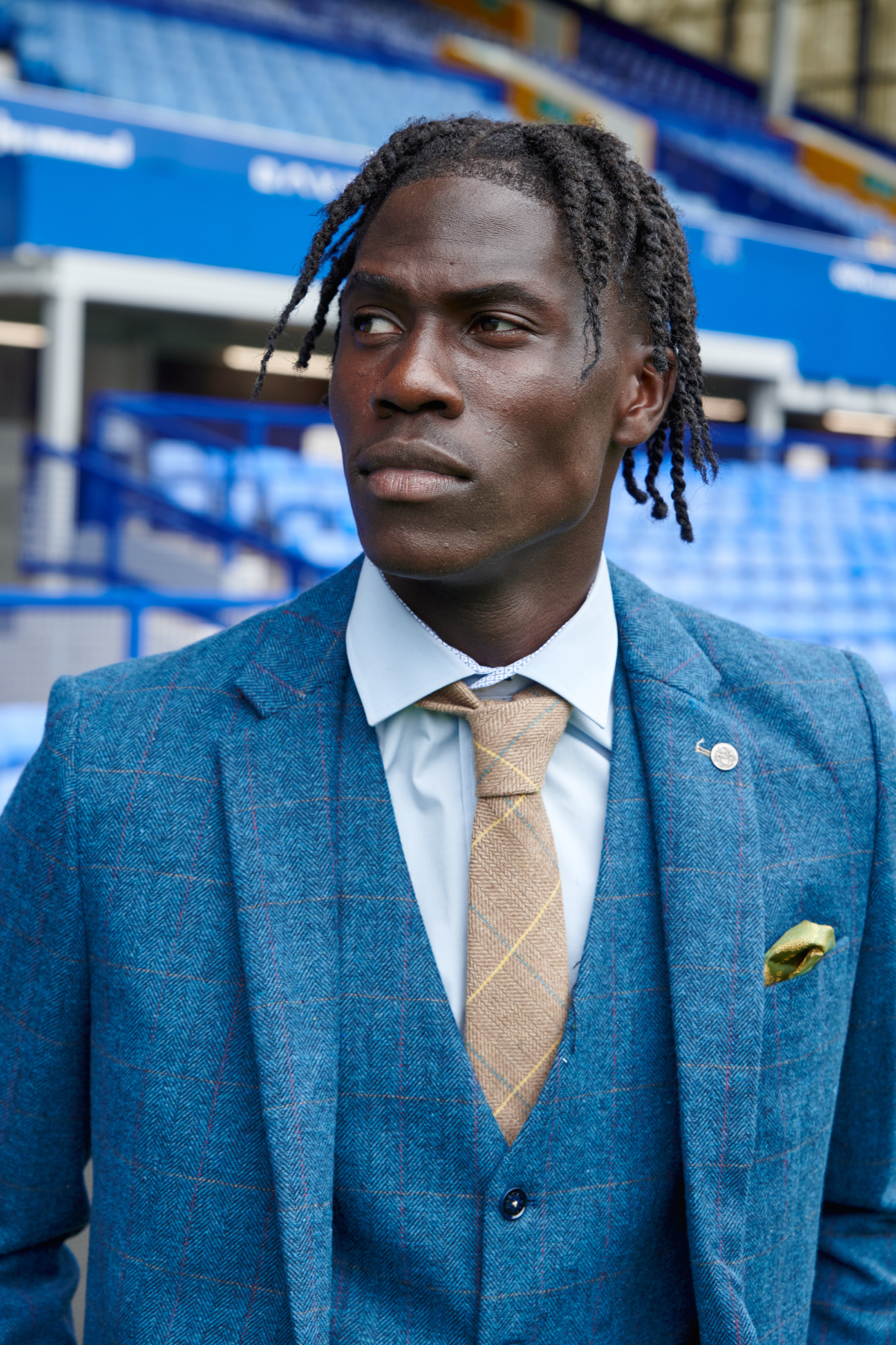 Amadou Onana wears the marcdarcy Everton Collection Dion Blue suit with vest and patterned tie, paired with a yellow pocket square. He stands in an empty stadium with blue seats and railings, gazing to the side.