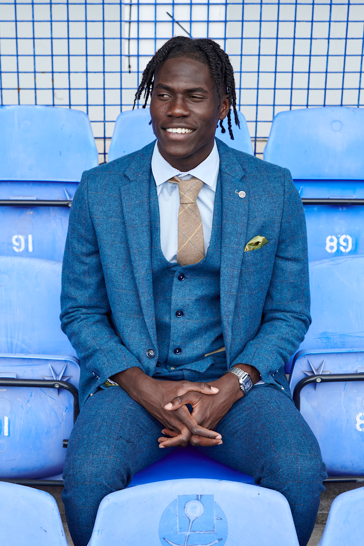 Amadou Onana wears marcdarcy's The Everton Collection | Dion Blue suit with a beige tie, white shirt, yellow pocket square, and watch. He smiles on light blue stadium seats with blue metal fencing in the background.
