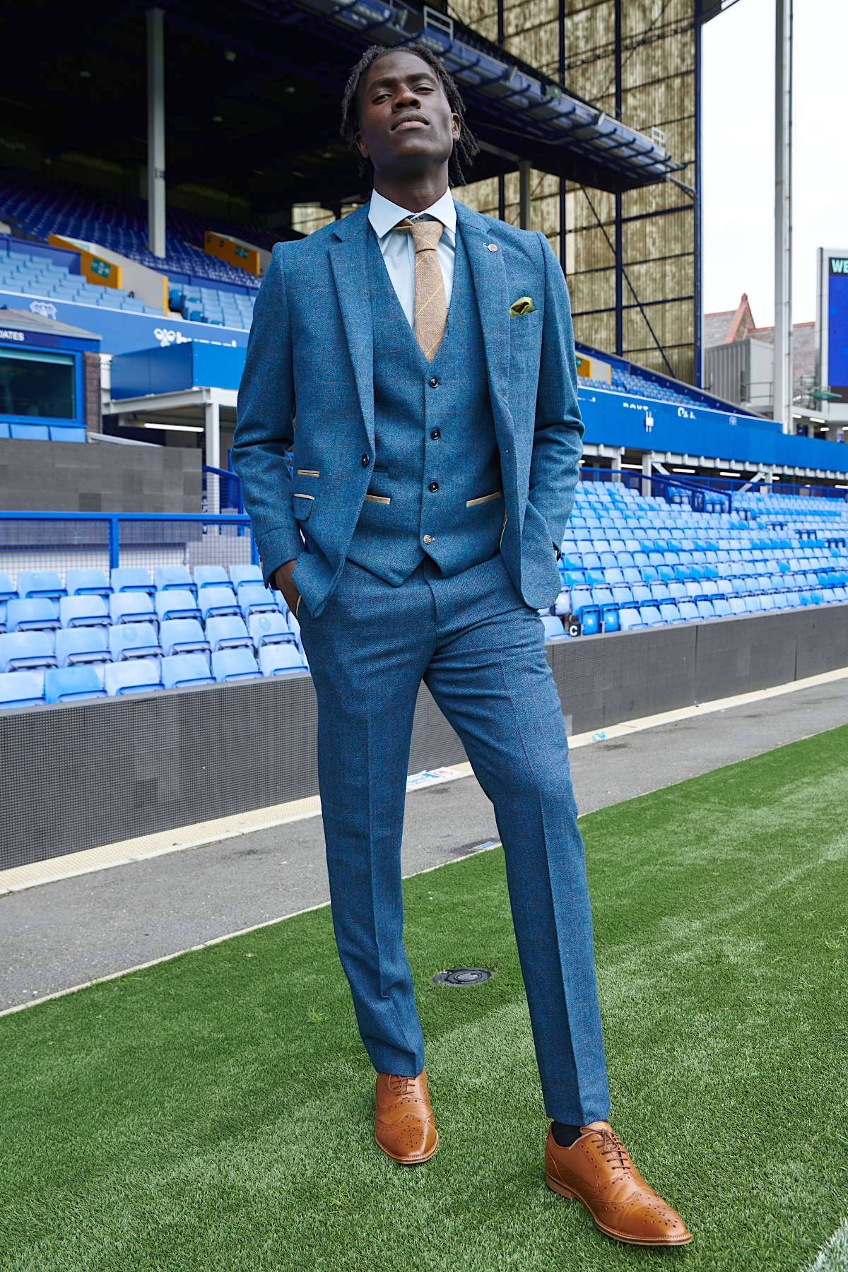 Amadou Onana models The Everton Collection by marcdarcy, wearing a Dion Blue suit with tan accents and brown dress shoes, exuding classic men's formal style on a football field with stadium seating in the background.