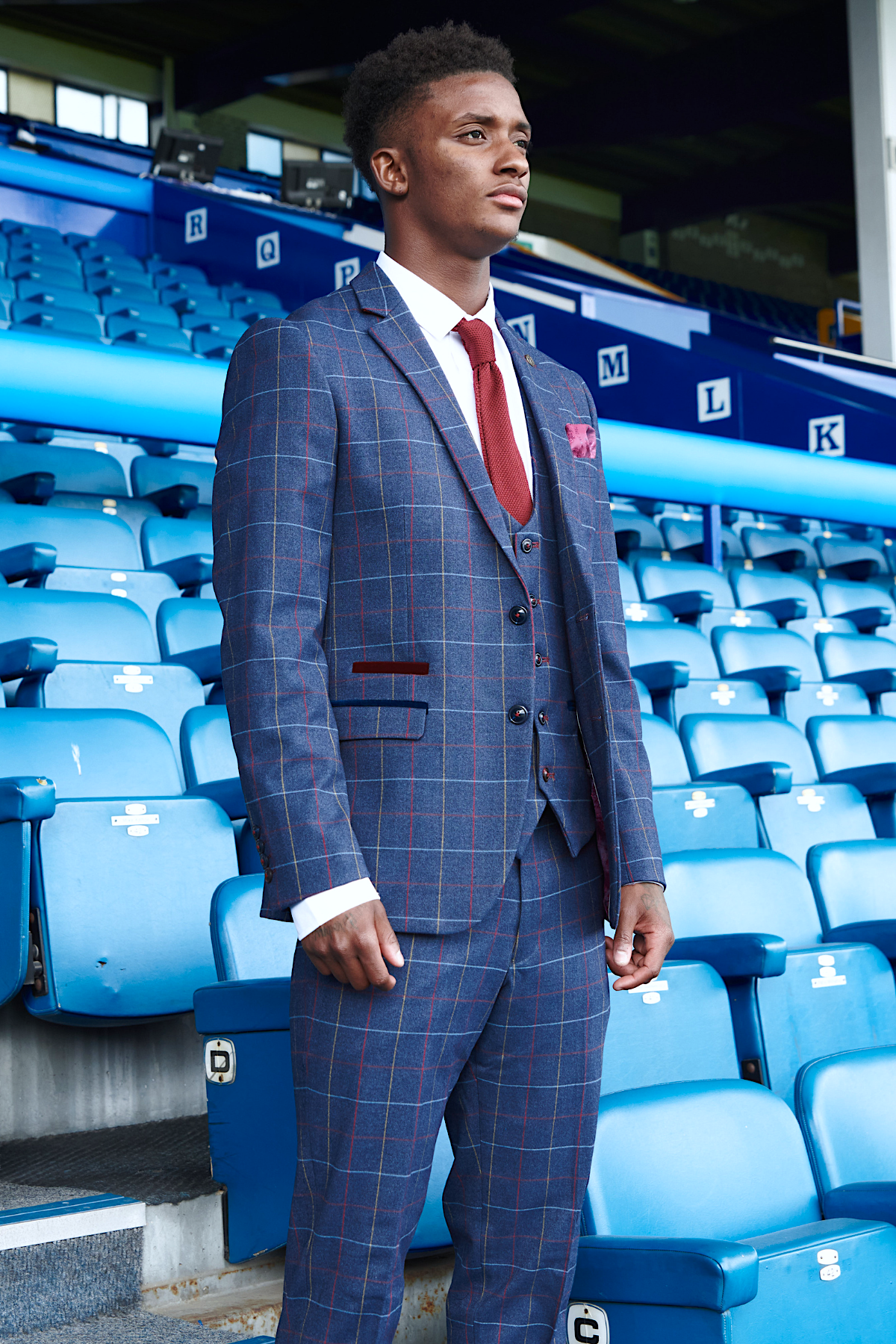 Demarai Gray models the marcdarcy Drake Navy Suit from The Everton Collection, standing confidently in a stadium with empty blue seats behind him.