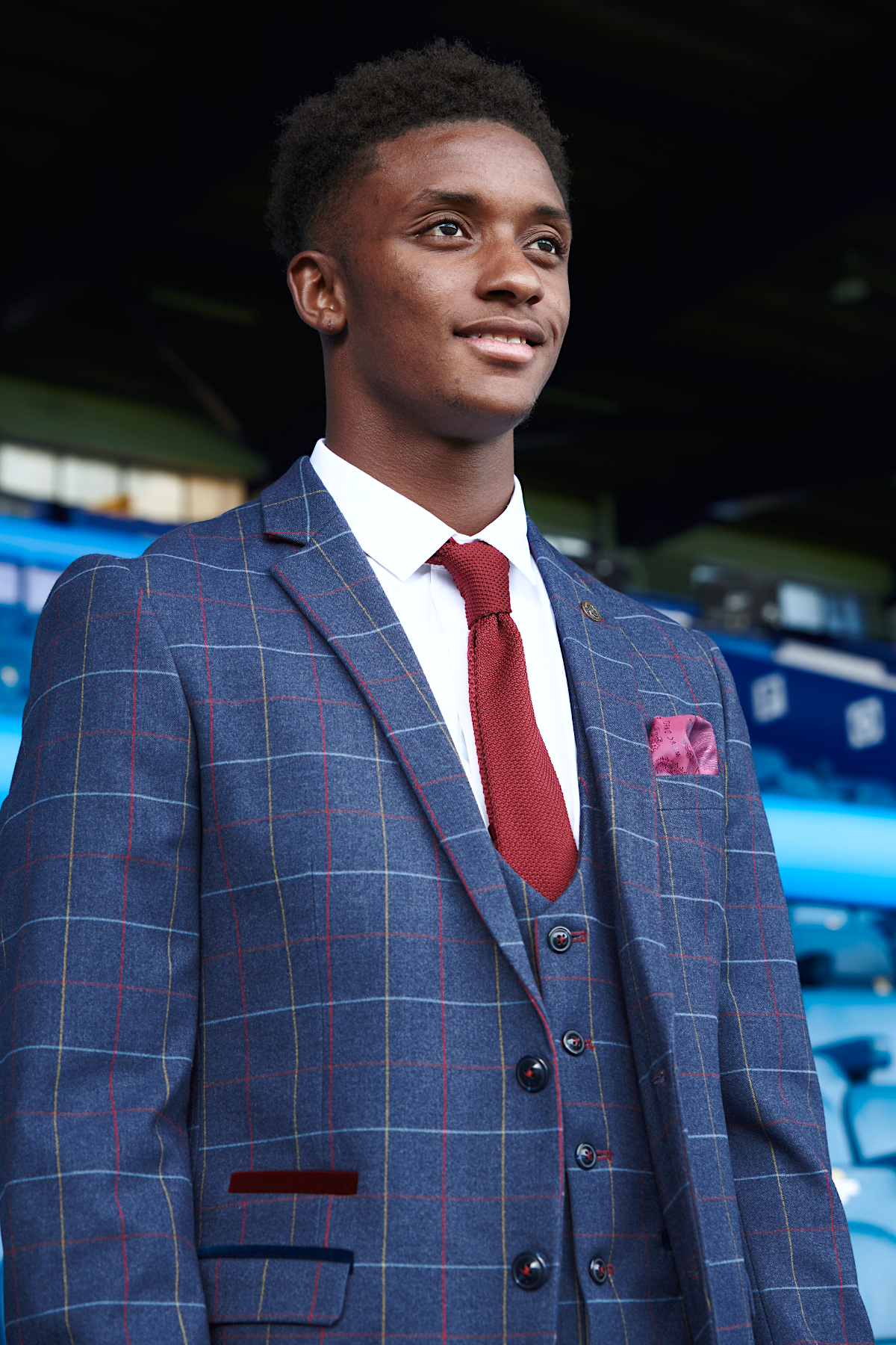 Demarai Gray stands confidently in a stadium with blue seats, wearing marcdarcy’s The Everton Collection Drake Navy Suit.