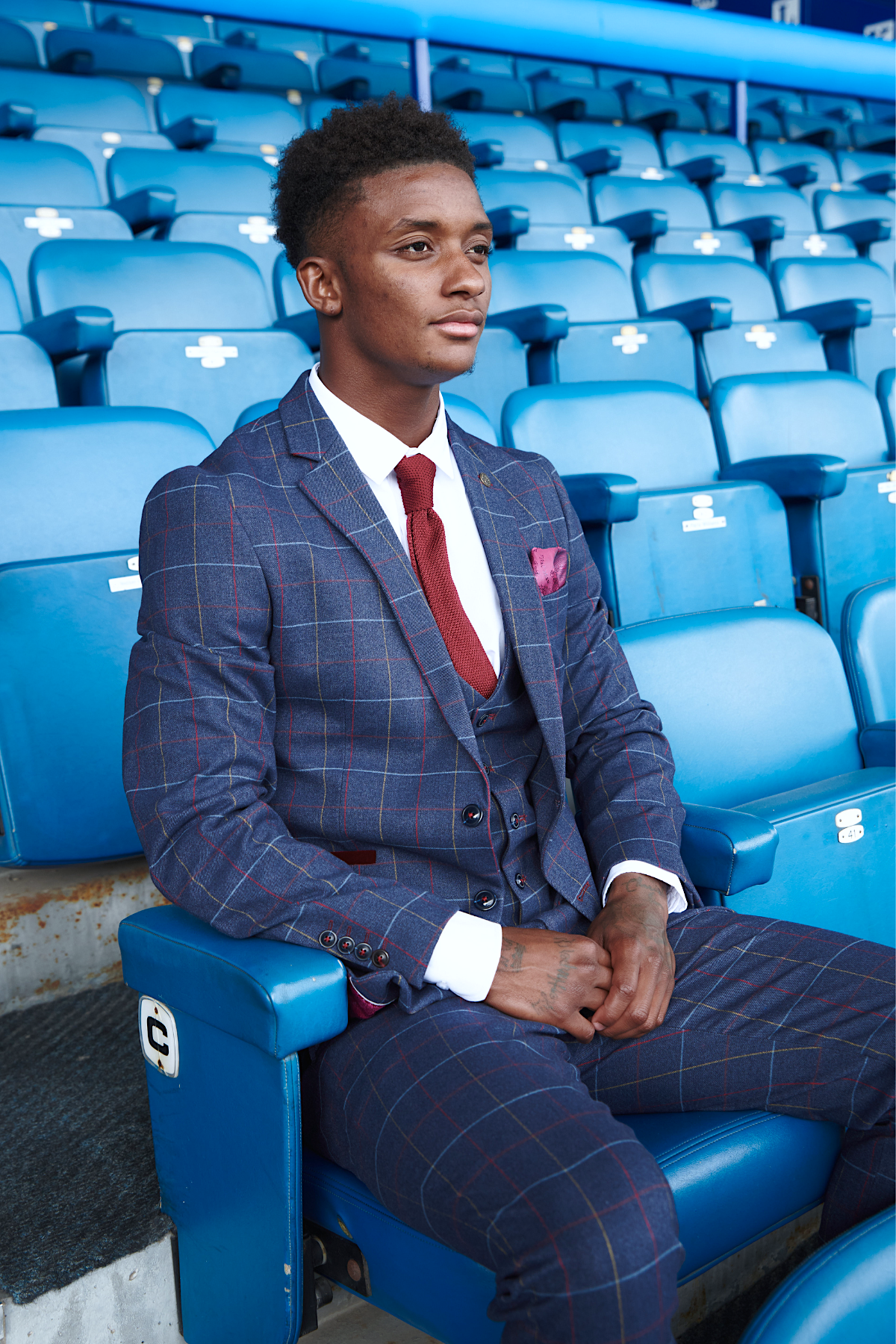 Demarai Gray from The Everton Collection by marcdarcy sits in an empty blue stadium seat, looking ahead calmly in a slim fit Drake Navy suit with a red tie and pocket square.