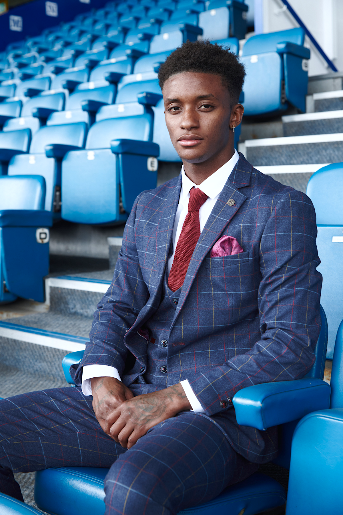 Demarai Gray models the marcdarcy Drake Navy Suit from The Everton Collection, sitting confidently in a blue stadium seat with empty rows and steps in the background.