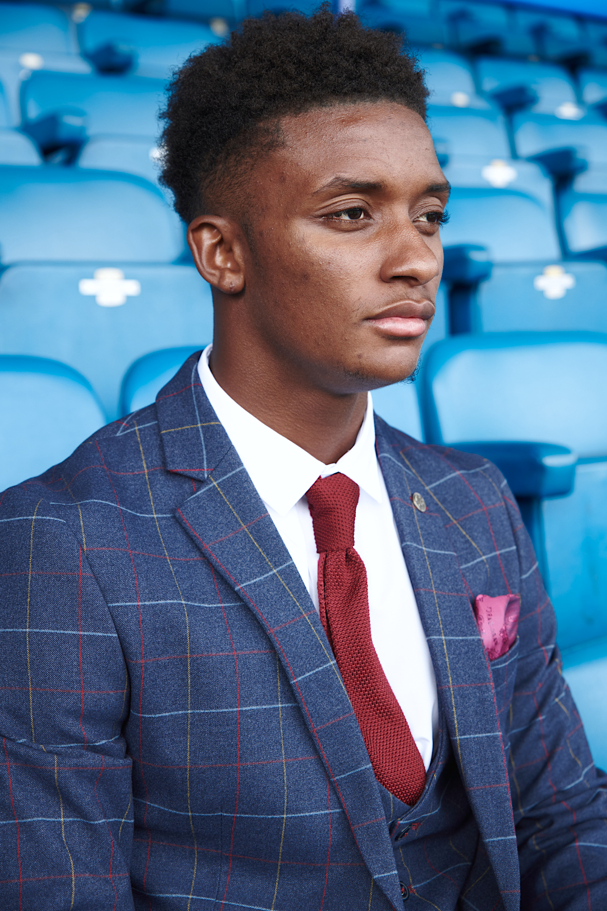 Demarai Gray wears a Drake Navy Suit from marcdarcy's The Everton Collection, paired with a red tie and white shirt, as he sits thoughtfully in a stadium with blue seats.