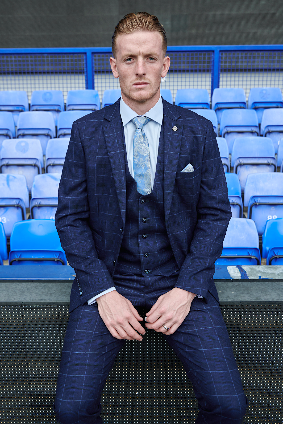 Jordan Pickford wears marcdarcy’s Edinson Navy Sky slim fit three-piece suit from The Everton Collection, sitting on a barrier before empty blue stadium seats, looking seriously at the camera.
