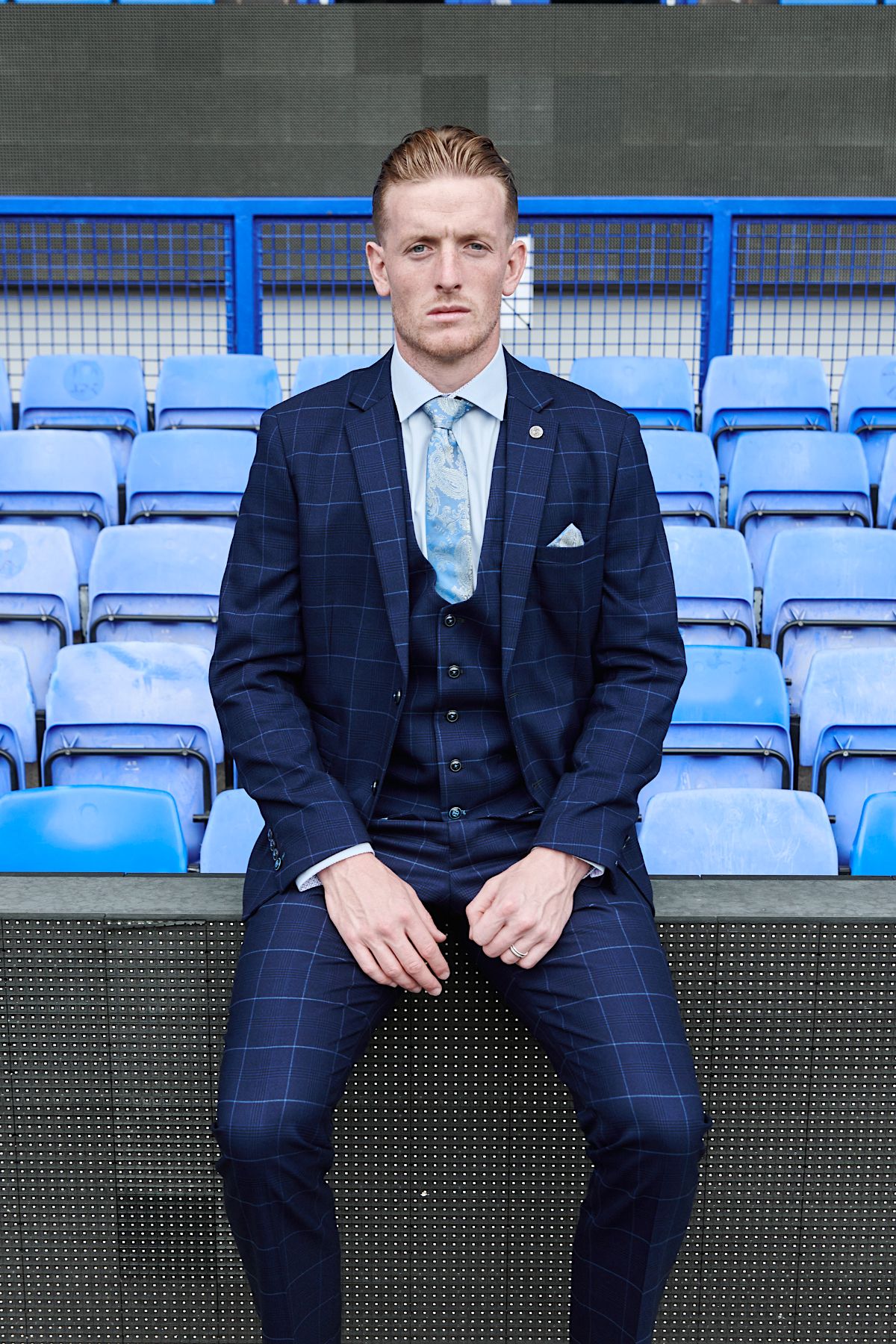 Jordan Pickford models The Everton Collection in an Edinson Navy Sky suit by marcdarcy, seated on a stadium bench with blue seats behind him.